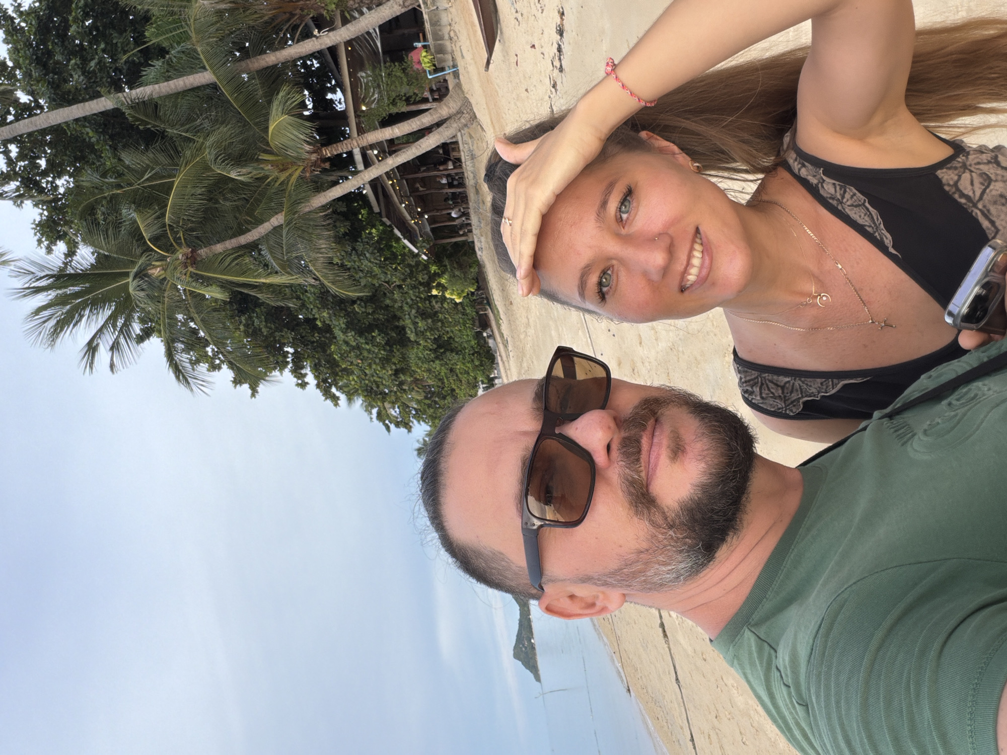 In the city of Baan Thong Po, Thailand, a man and a woman are taking a selfie on a beach. The man, on the left, is wearing a green t-shirt and sunglasses, with his hair tied back. The woman, on the right, is wearing a black top with a patterned detail and multiple necklaces. She is smiling and touching her forehead, with her hair pulled back. Behind them, palm trees and a rustic wooden structure, possibly a restaurant or bar, are visible. The beach sand is light brown, and the ocean is in the background, with a distant island or landmass. The sky is overcast.