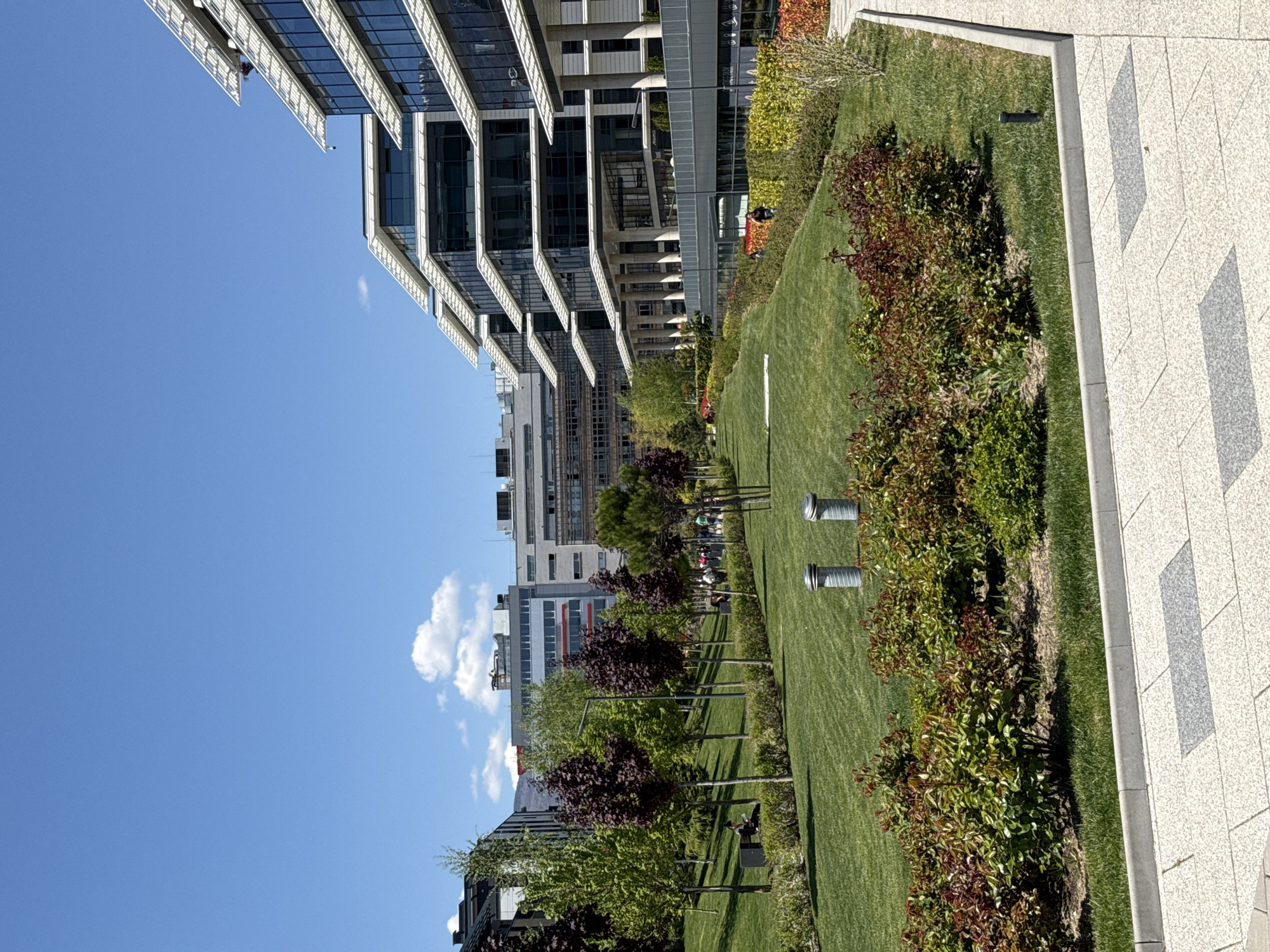 The image shows a park area with a lawn, bushes, and trees adjacent to modern office buildings. The scene is likely located in Sofia, Bulgaria. On the right side of the frame, there is a tiled walkway, and the grass area contains several lighting fixtures. People are visible sitting on benches in the park. The background includes several buildings of various heights under a blue sky with a few clouds. The perspective is taken at ground level, with a tilt towards the left.