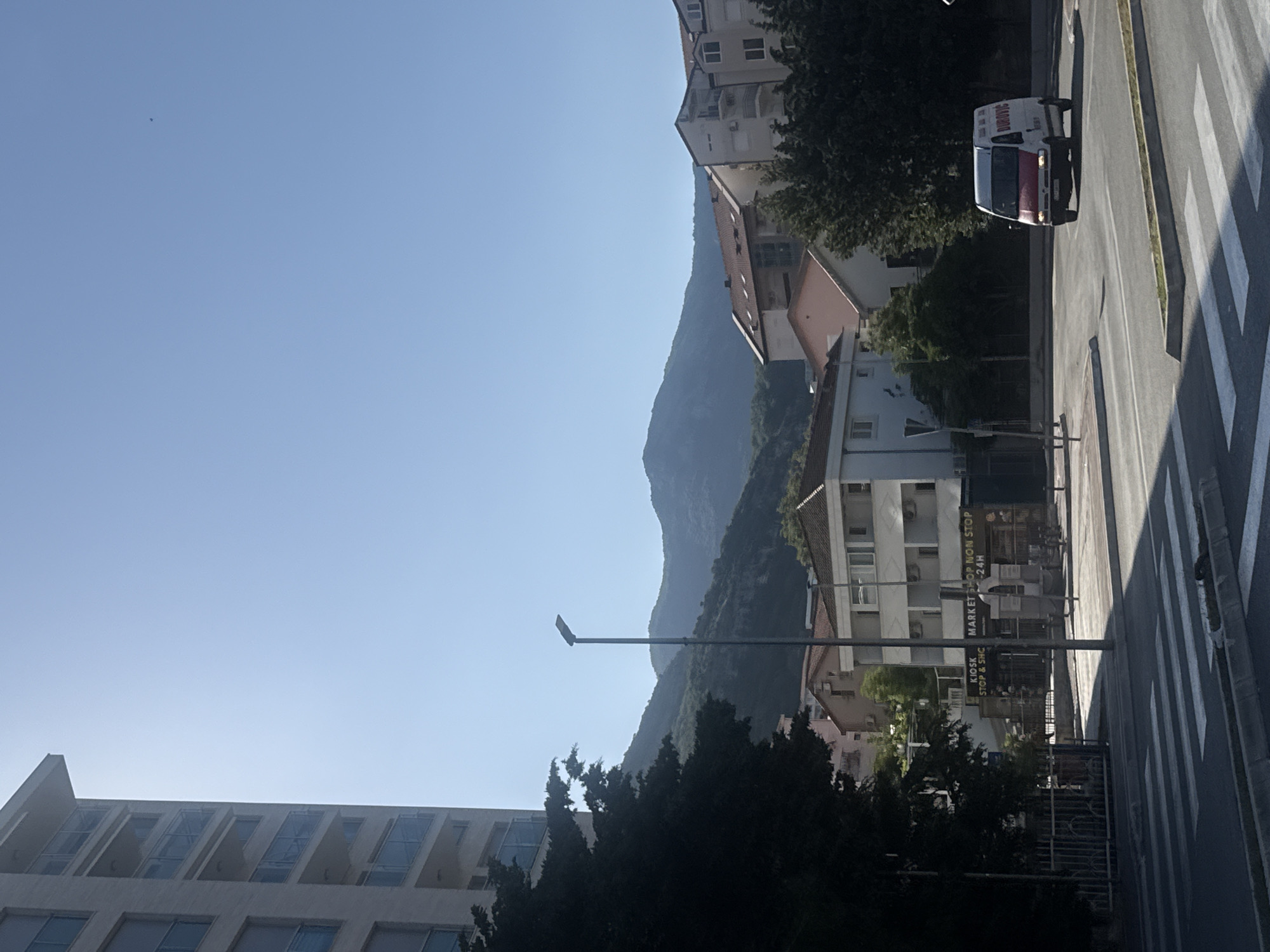 The image depicts a sunlit urban street scene in Rafailovići, Montenegro, with mountains in the background. The sky is clear and blue, with a single faint dark speck, possibly a distant bird or aircraft, visible in the upper right quadrant.
In the immediate foreground, a white-striped zebra crossing is marked on dark asphalt pavement, which transitions into a concrete curb and a narrow, raised median strip. A tall, dark grey streetlamp pole stands centrally in the mid-left, extending above the treeline.
To the left, a multi-story modern building with light-colored facade panels and glass-fronted balconies rises prominently. Partially obscuring its lower section is a dense, dark green tree, casting a deep shadow. Further into the midground, beyond the tree and streetlamp, various residential and commercial structures are visible. A building with storefronts displays signs, with legible portions reading "KIOSK STOP & SHC" and "MARKET SHOP NON STOP 24H". Other buildings feature reddish-brown tiled roofs and multiple windows, some with external air conditioning units.
On the right side of the road, a white van with a red lower section is in motion, appearing to turn left. The word "ĐUROVIC" is partially visible in dark lettering on its side. More trees and residential buildings are situated behind the van and to its right.
The background is dominated by a range of verdant, rugged mountains under a bright, cloudless sky, indicating strong sunlight and clear visibility. Shadows are distinct, suggesting a time of day with high sun. No pedestrians are clearly visible.