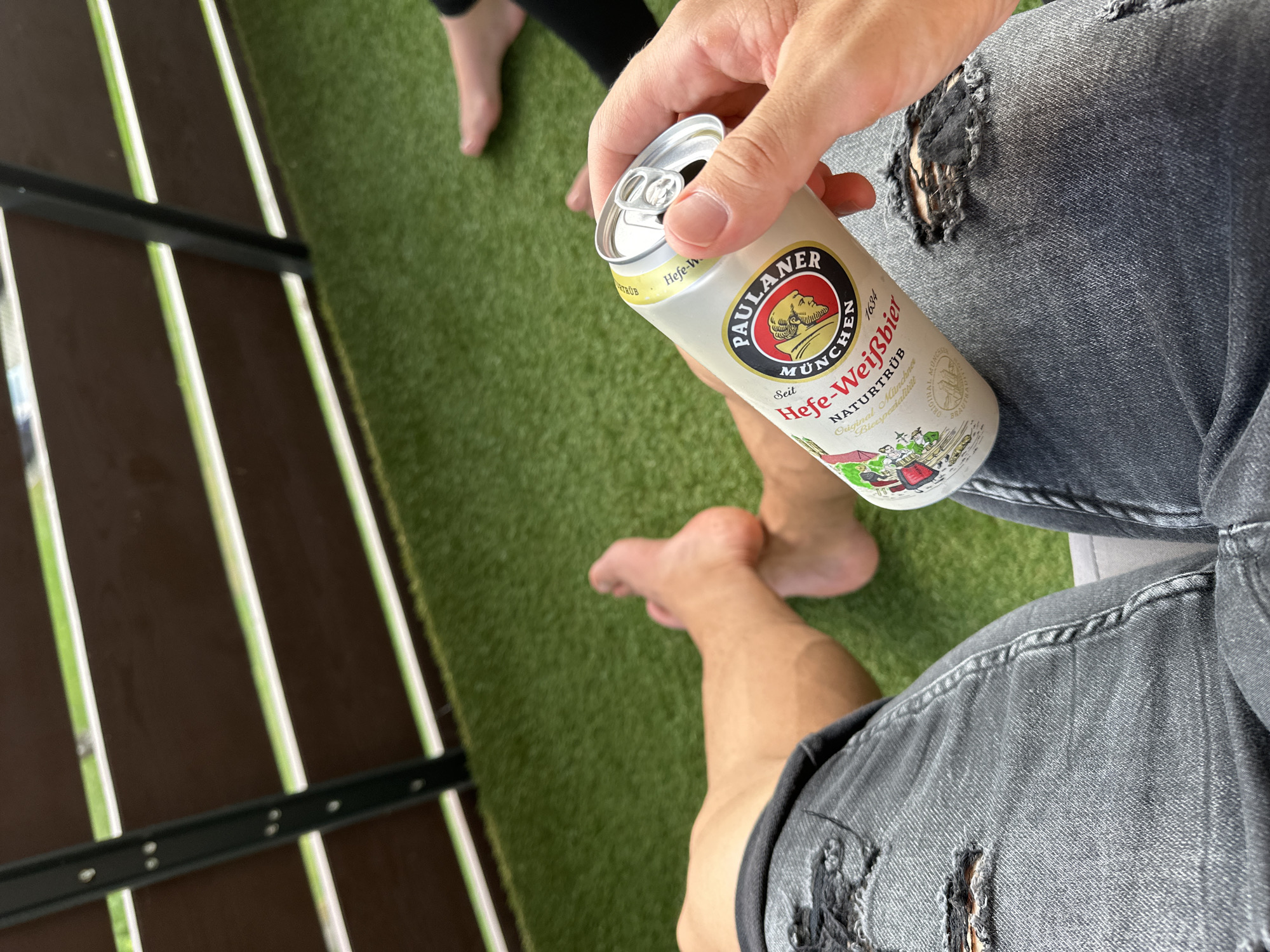 The image shows a person's legs and hand holding a can of Paulaner Hefe-Weißbier. The person is sitting on a chair, with artificial grass flooring and a wooden railing in the background. The overall scene suggests a casual outdoor setting, possibly a balcony or patio. It is impossible to determine the exact location within Garmisch-Partenkirchen, Germany based solely on the visual information provided.