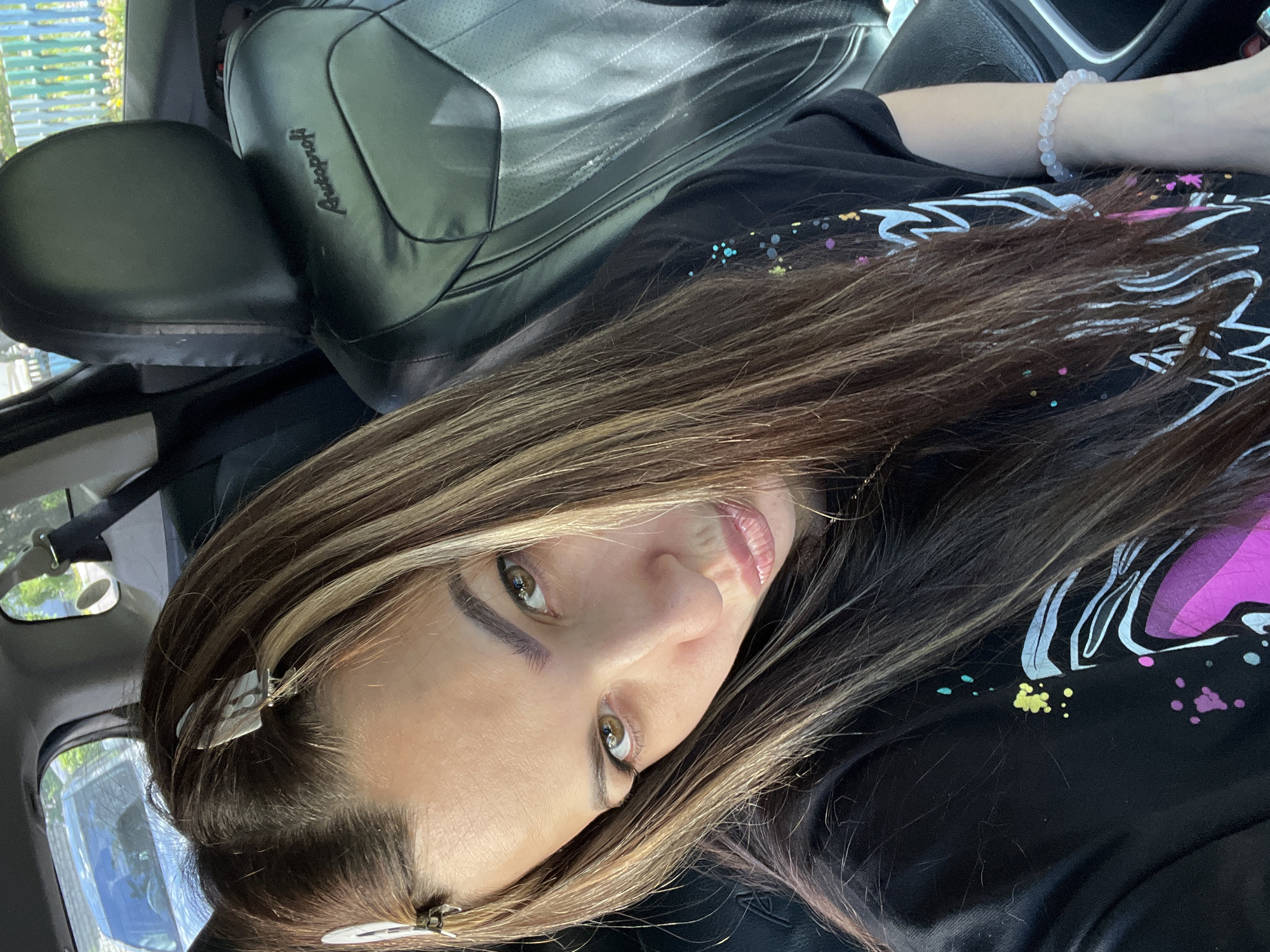 A woman is in a car, presumably the driver's seat, in Unknown, Unknown. She has long brown and blonde hair. Her hair is styled with hair clips. She is wearing a black shirt with a colorful design. She also has a bracelet of pinkish beads on her wrist. The car seat is black. The environment is daytime, with sunlight visible through the car window.