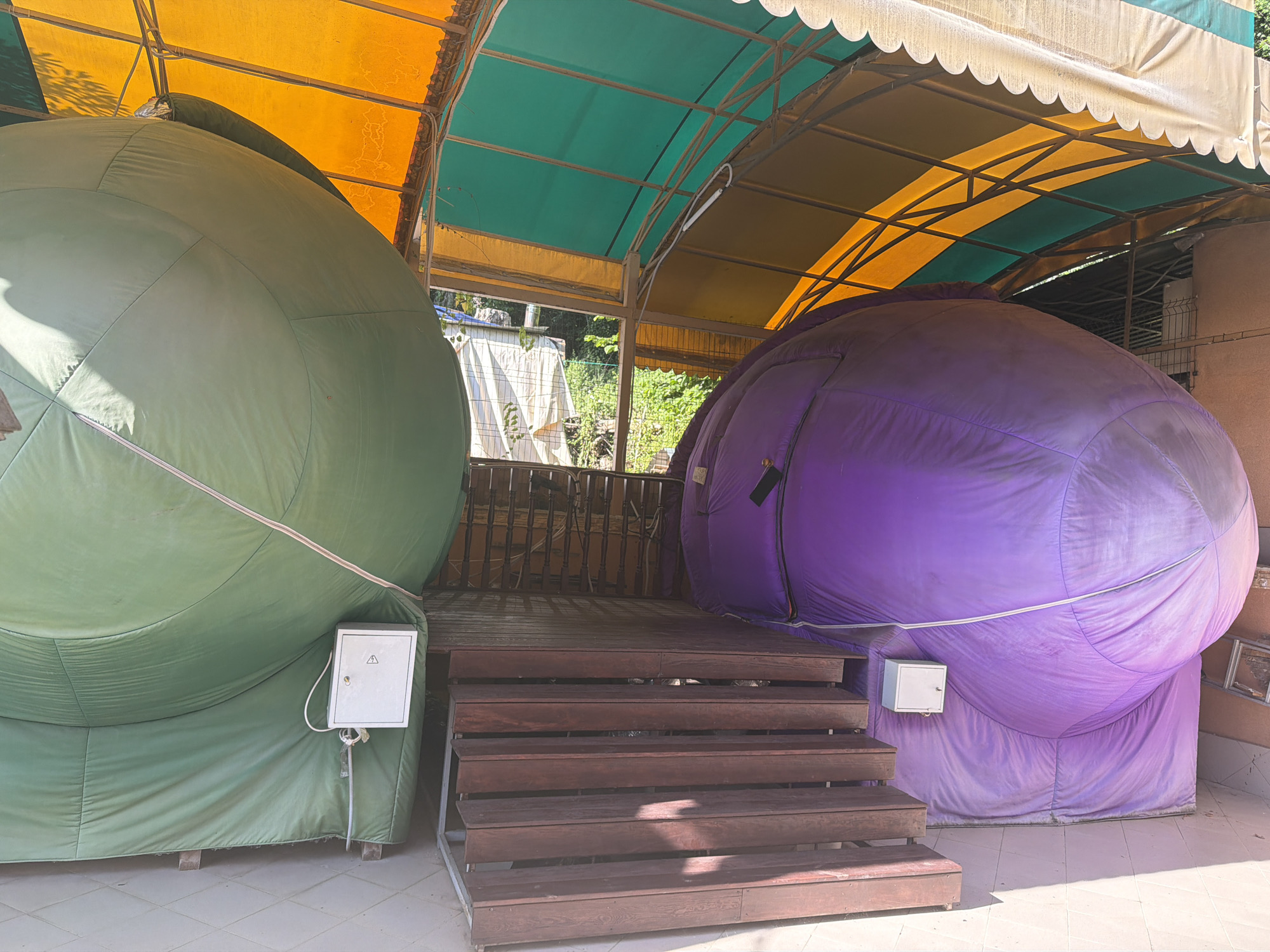 This media file shows two large inflatable structures, one green and one purple, situated outdoors under a colorful awning. The awning is made of alternating yellow and green fabric panels supported by a metal frame, with a scalloped white trim along the edge. The structures appear to be dome-shaped and are resting on wooden bases.
In front of the inflatables is a wooden deck with a set of wooden stairs leading up to it. Each inflatable has a small white electrical box mounted on its side, with one box visible on the green inflatable and another on the purple inflatable. The green inflatable has a white triangular symbol on its electrical box, indicating a potential hazard.
The scene is set outdoors, likely during the daytime, given the bright sunlight casting shadows. The background shows greenery and some fencing, suggesting a possibly recreational or park-like setting. The overall impression is of a playful or whimsical installation, perhaps for an event or attraction. There is no visible text other than the warning symbol. The location is identified as Sochi, Russia. No people are visible in the image. The weather appears to be clear and sunny.