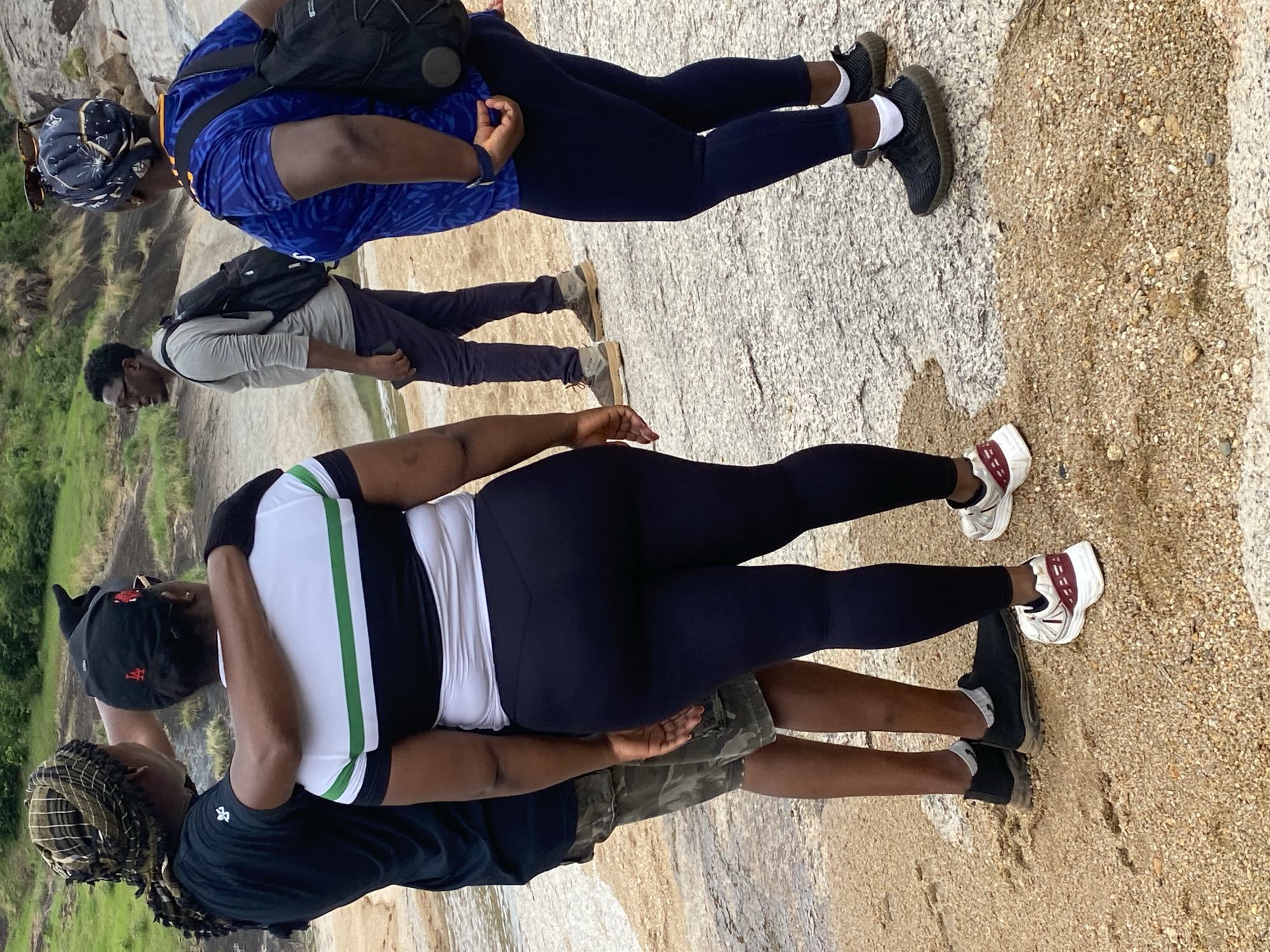 The image shows a group of four people outdoors, seemingly on a rocky surface. The location appears to be Ganawuri, Nigeria.
A person wearing a blue patterned shirt and black leggings stands at the top with a backpack. Below them, a person in a grey long-sleeved shirt and dark pants stands as well with a backpack. Further down, a person wearing a black and white shirt and black leggings stands next to a person wearing a black shirt and camouflage shorts. The image is taken at an angle, looking down on the group. The ground is a mix of rocky and sandy terrain. The background consists of trees and possibly another rock formation. The people are interacting and appear to be posing for the photo.