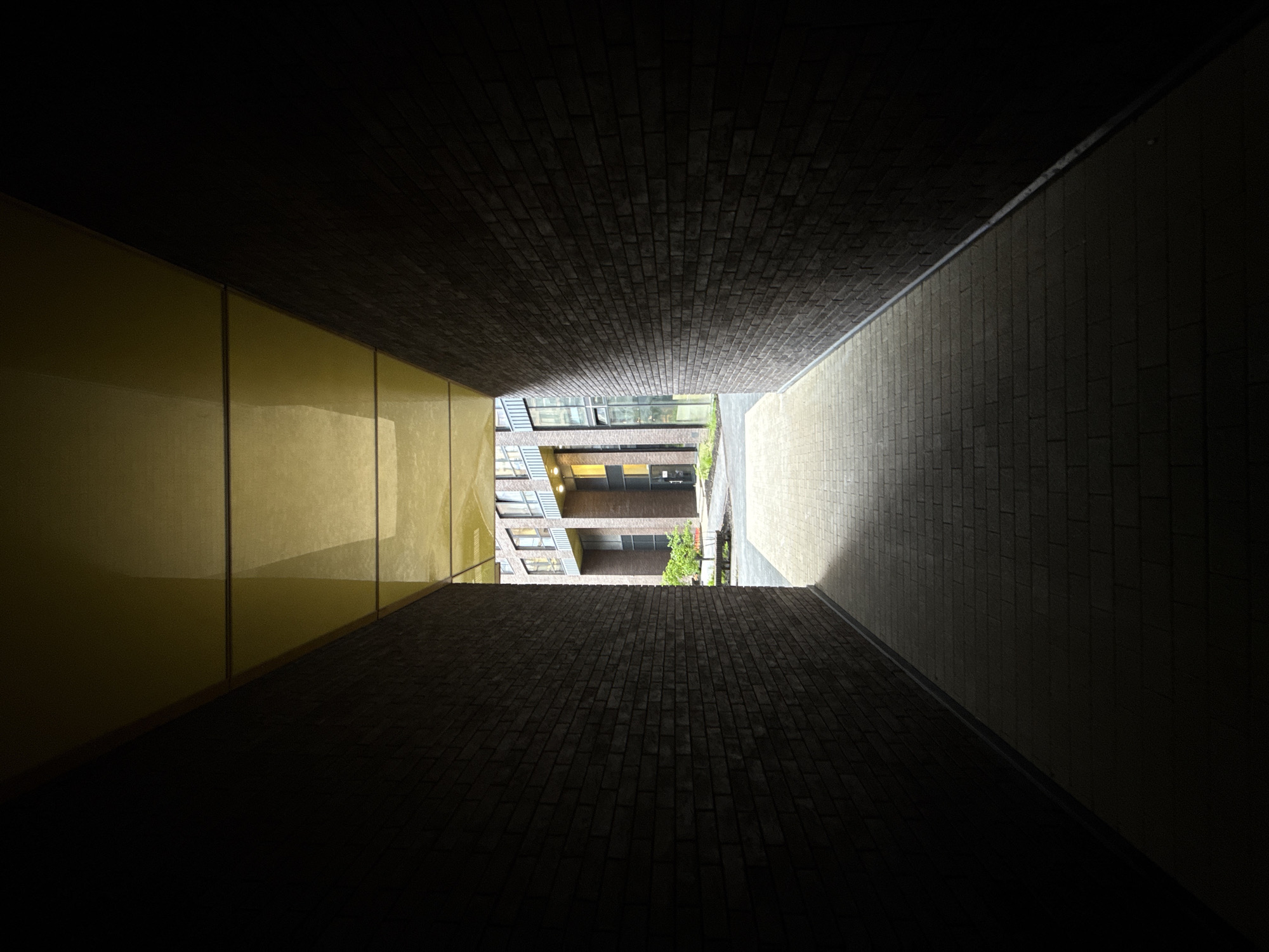 The image depicts a narrow passageway in an urban setting, looking outward towards an open courtyard. The walls of the passageway are constructed of dark brick on one side and a light yellow, segmented material on the other. The passageway appears dark, with light increasing towards the opening at the far end. The floor is paved with light-colored bricks. Beyond the passageway is a courtyard with buildings, foliage, and a patch of sunlight. The buildings have windows and a brick facade. There are no people visible in the image. The location is Moscow, Russia.