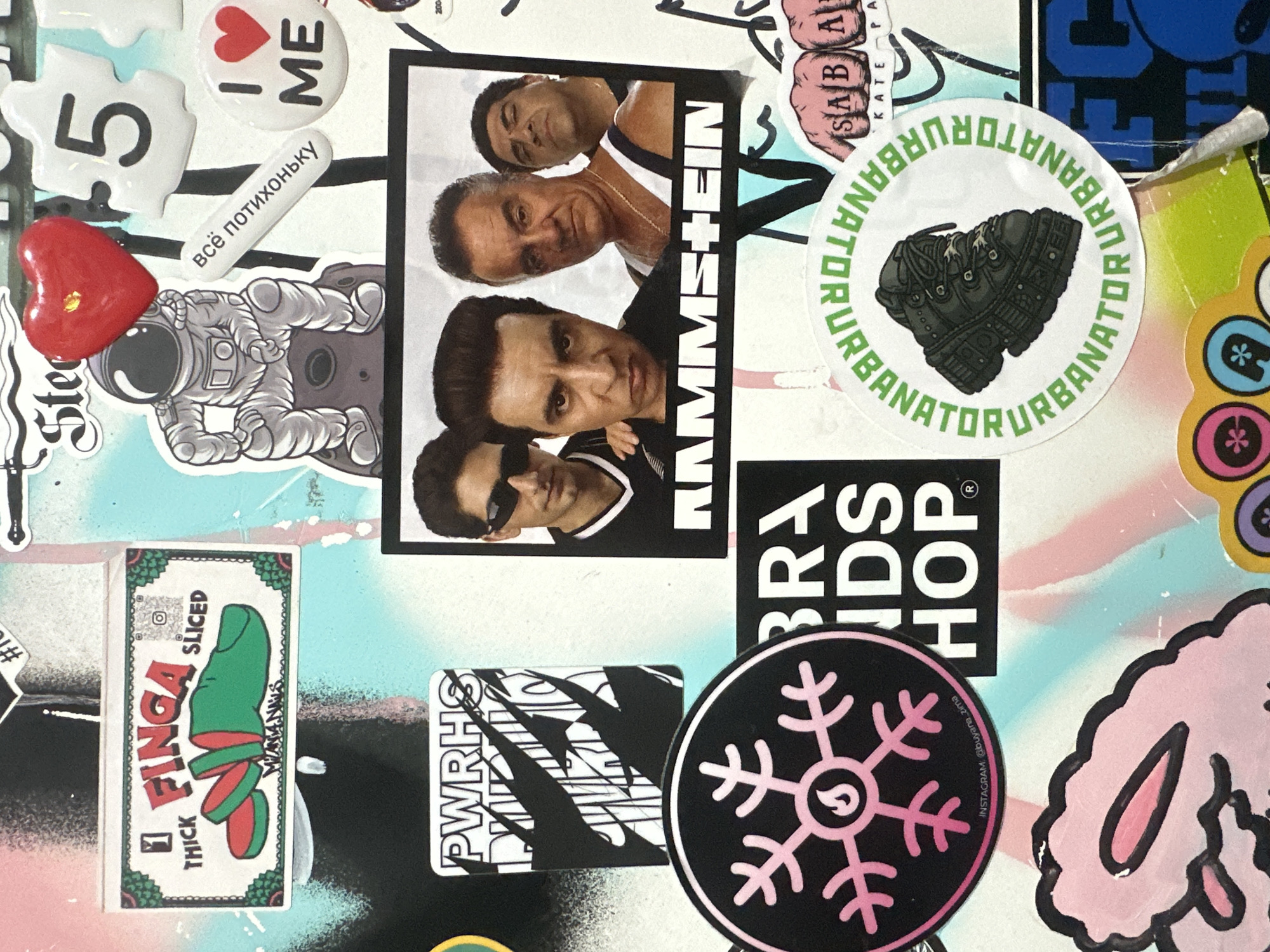 This media file is a collage of various stickers and decals applied to a surface, likely a wall or board, with a spray-painted background featuring abstract designs and colors like blue, pink, and white. The collage includes several distinct elements.
Prominently featured is a black and white photograph of four men, likely from a band, with the text "RAMMSTEIN" vertically oriented alongside them. Another circular sticker displays a close-up illustration of a black boot with the text "URBANATOR" repeated around it.
There are also several smaller stickers. One is a red heart with gold flecks. Another is an astronaut figure. A white oval sticker has a heart symbol and the text "I ME". A white rectangular sticker with a green border depicts a sliced finger, labeled "FINGA", with the words "THICK" and "SLICED". There's also a black circular sticker with a pink snowflake-like design and a flame symbol in the center.
Visible text includes Russian phrases like "всё потихоньку" (everything in due time), and English text such as "PWRHS", "BRA IDS HOP", and Instagram handles. The overall impression is one of a collection of personal interests, possibly reflecting music tastes, humor, and individual style, applied as decorations in what could be a personal workspace or a public area in Moscow, Russia, given the location context. The scene is static, with no activity or event taking place other than the display of the stickers themselves.