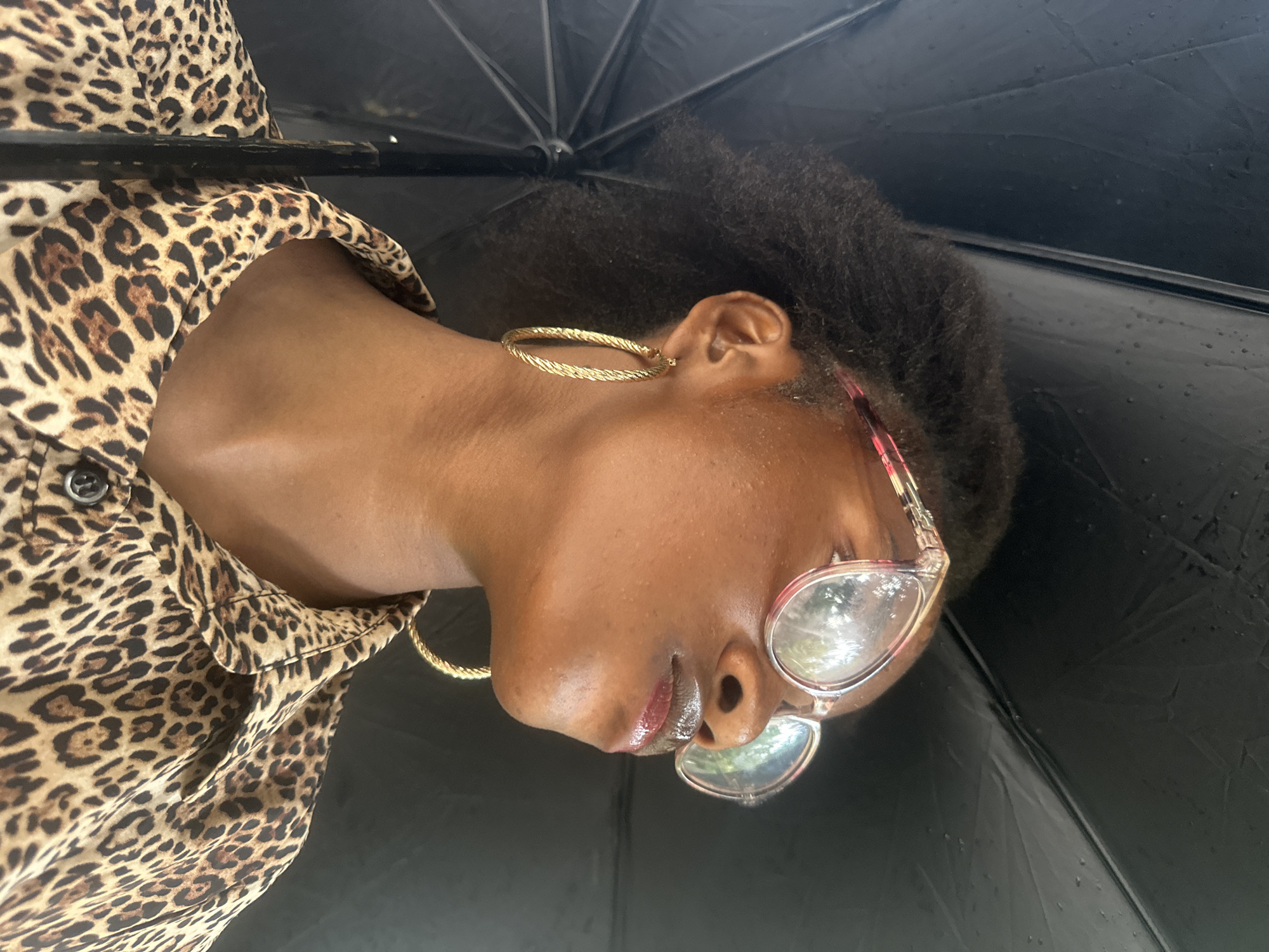 The image shows a close-up of a young Black woman from the shoulders up, looking to her right. She is under a black umbrella, which takes up a significant portion of the background. The woman has her hair pulled back, and she is wearing large, clear-rimmed glasses with a pinkish hue. She has large gold hoop earrings and is wearing a leopard-print button-down shirt. The scene appears to be outdoors, possibly in the city of Badagry, Nigeria, given the context provided. There are small water droplets visible on the umbrella, suggesting it is raining or has recently rained. The lighting is soft, with reflections visible on her glasses and lips.