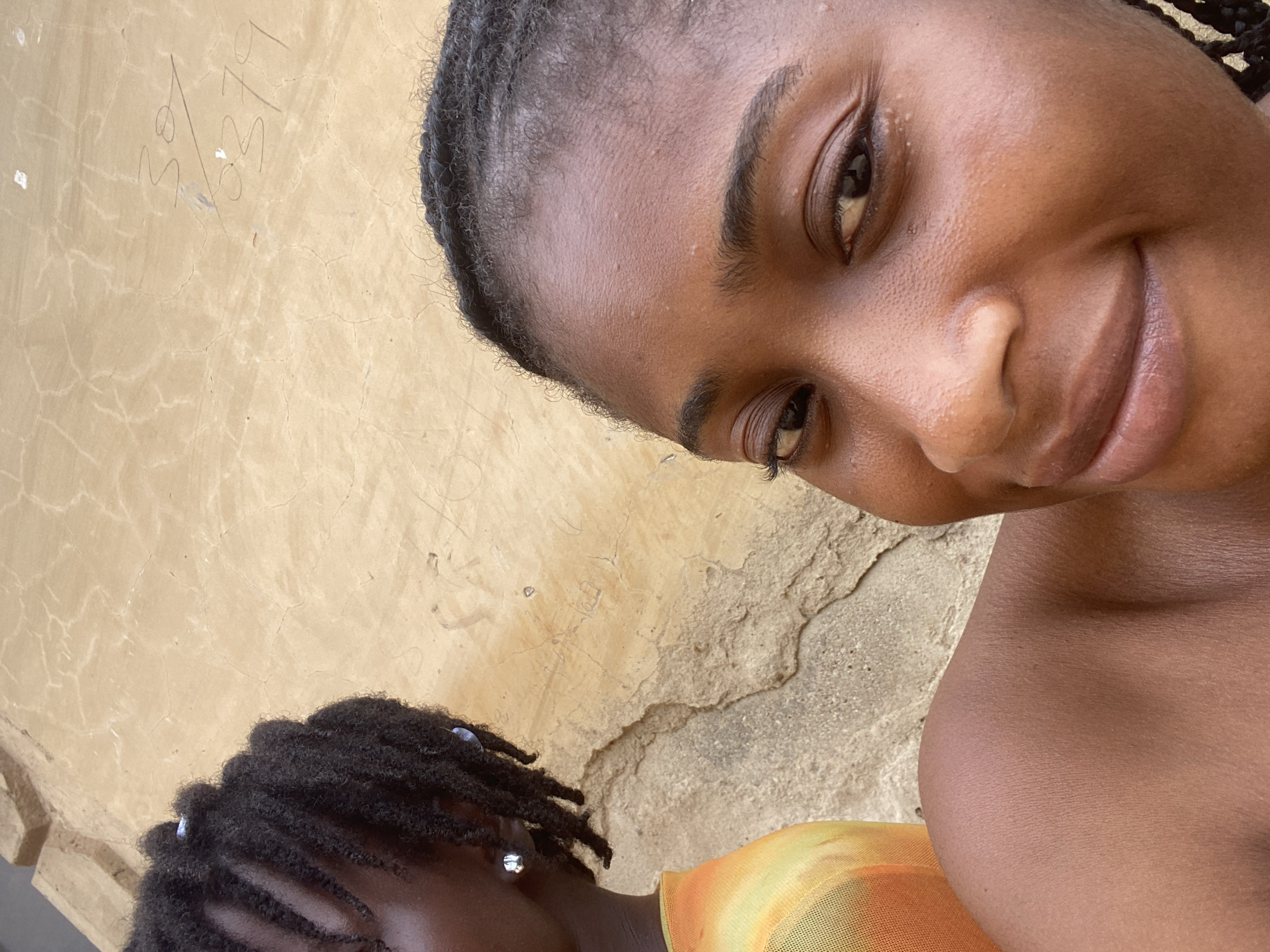 The scene depicts two women in Kubwa, Nigeria, positioned against a textured, light brown wall. The woman in the foreground, on the right, has dark skin and is visible from the chest up, looking directly at the camera with a soft smile; her right shoulder and collarbone are bare, and her dark hair is closely styled on her visible forehead and scalp. The second woman, partially visible in the background on the left, also has dark skin with dark dreadlocks or twists, and wears an orange and yellow patterned top and a silver stud earring in her left ear. The background wall exhibits numerous hairline cracks and features the handwritten numbers "30 / 379" in dark pigment in the upper right quadrant. A rougher, darker concrete surface is visible below the main wall. The environment is illuminated by natural daylight.