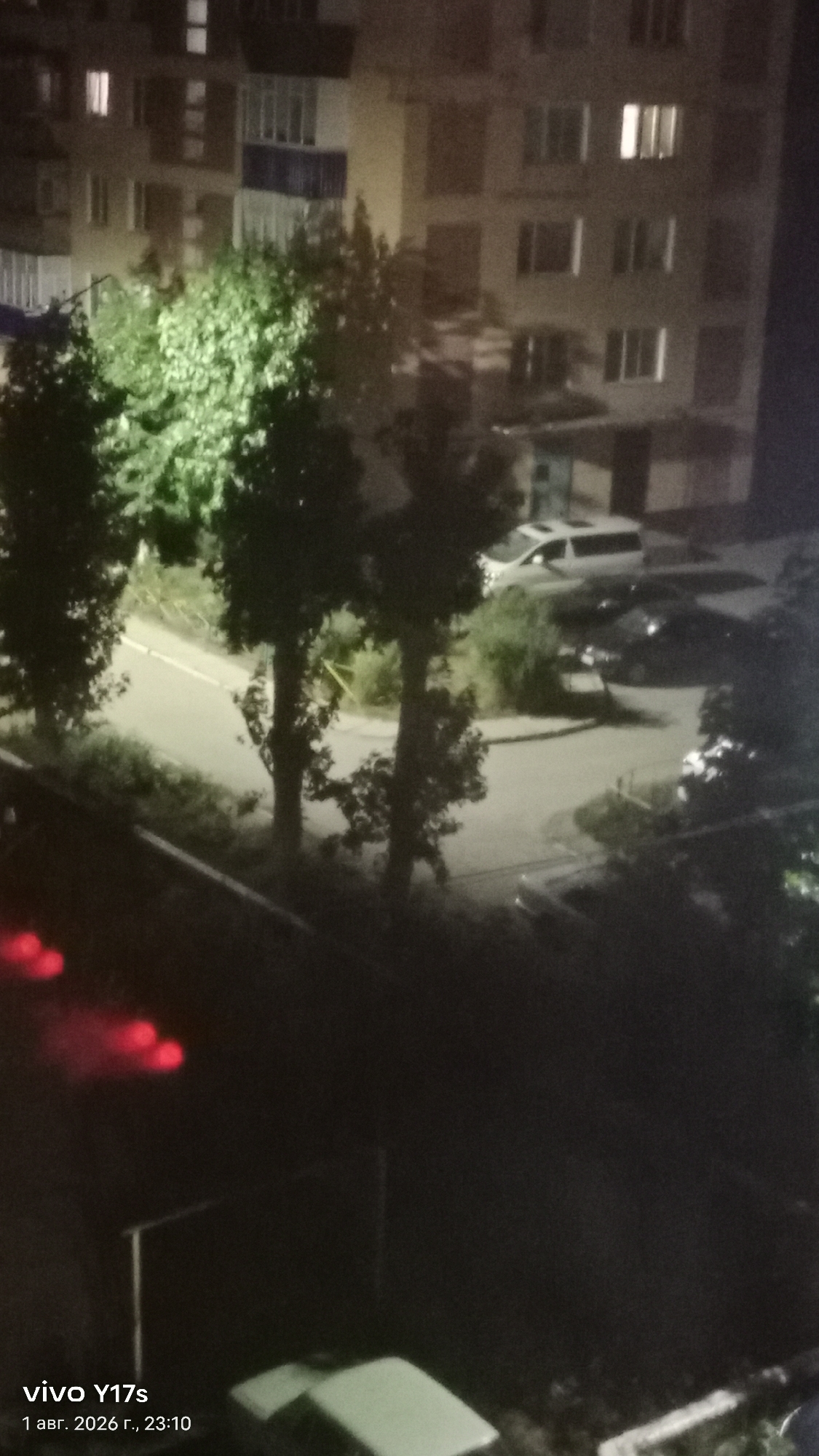 The image is a nighttime scene looking down at a street with trees and apartment buildings in the background. A white van and several cars are parked on the street. The lighting suggests artificial illumination from streetlights or building windows. The bottom left corner shows a watermark that reads "vivo Y17s 1 авг. 2026 г., 23:10". This indicates the photo was taken on August 1, 2026, at 11:10 PM, with a vivo Y17s phone. The date format suggests the location might be Russia or a country using a similar calendar system. The city of Rudny, Kazakhstan, is a known location for a similar date format.