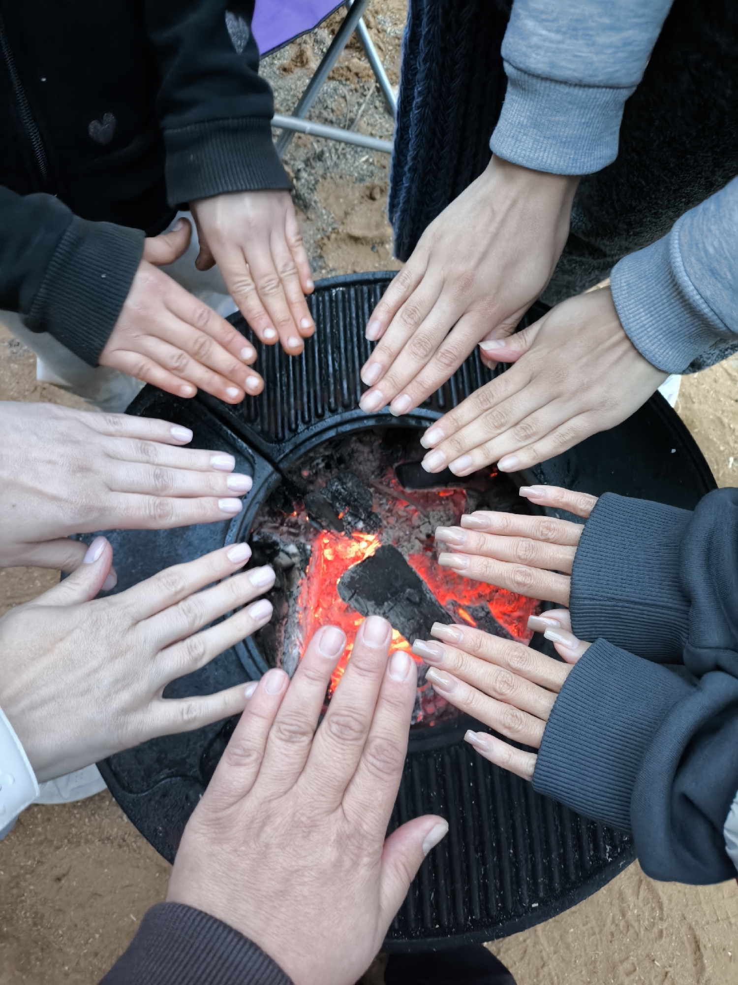 A high-angle, full-frame shot depicts a group of people surrounding a fire pit, warming their hands over glowing embers. The fire pit is black and appears to be made of cast iron, with a grate-like top. Around the pit, multiple hands are positioned close to the heat, suggesting an effort to warm themselves. The hands vary in skin tone and size, with some displaying manicured nails. The individuals are wearing various types of clothing, including a black hoodie with heart detailing, a light grey sweatshirt, a dark blue knitted scarf, a white sleeved shirt, and a dark brown jacket. A purple camping chair is partially visible in the background. The ground surrounding the fire pit appears to be sand or dirt. The location is Unknown, Unknown.