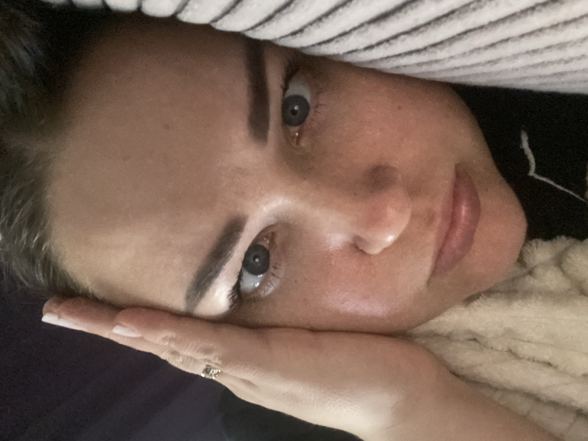 A close-up, rotated image shows a woman's face. Her hand rests near her cheek. She has light skin, dark eyebrows, and grey eyes. The background includes a striped fabric and a softer, textured material near her shoulder. The location is not discernible from the image.