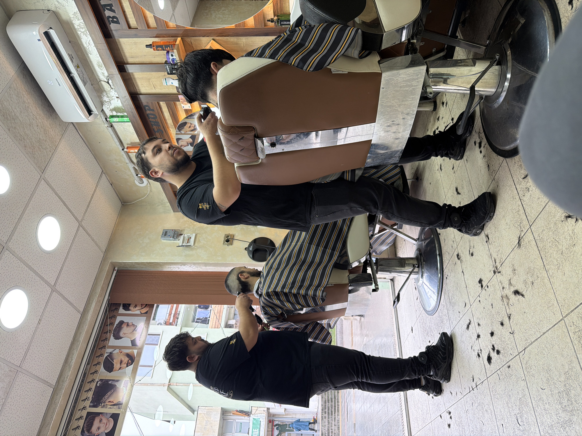 Inside a barber shop in Panagyurishte, Bulgaria, two barbers are cutting the hair of two customers. The barbers are both wearing black shirts and jeans. The customers are seated in barber chairs and are draped with striped barber capes. The floor is covered in hair clippings. The shop has a tiled floor and ceiling with fluorescent lighting. There is an air conditioning unit on the wall. Outside the window, a street scene is visible. There are pictures of different hairstyles on the wall.