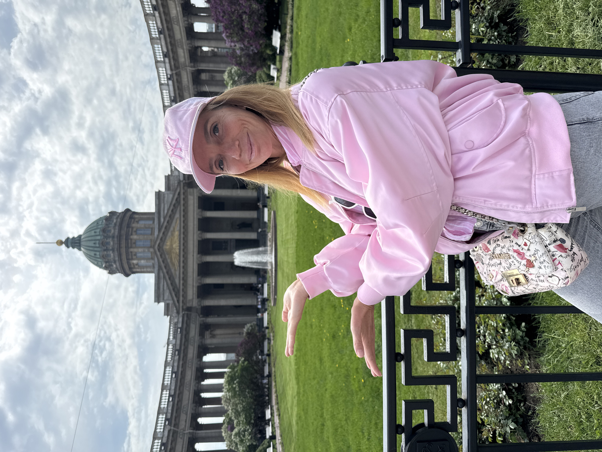 The image captures a woman in a pink outfit, leaning against a black metal fence. In the background is the Kazan Cathedral in Saint Petersburg, Russia, framed against a cloudy sky. The foreground includes green grass, trees, and a view of the Cathedral's façade and dome. The woman wears a pink jacket and cap and has a small patterned purse hanging from her shoulder. Her hands are resting on the fence.
