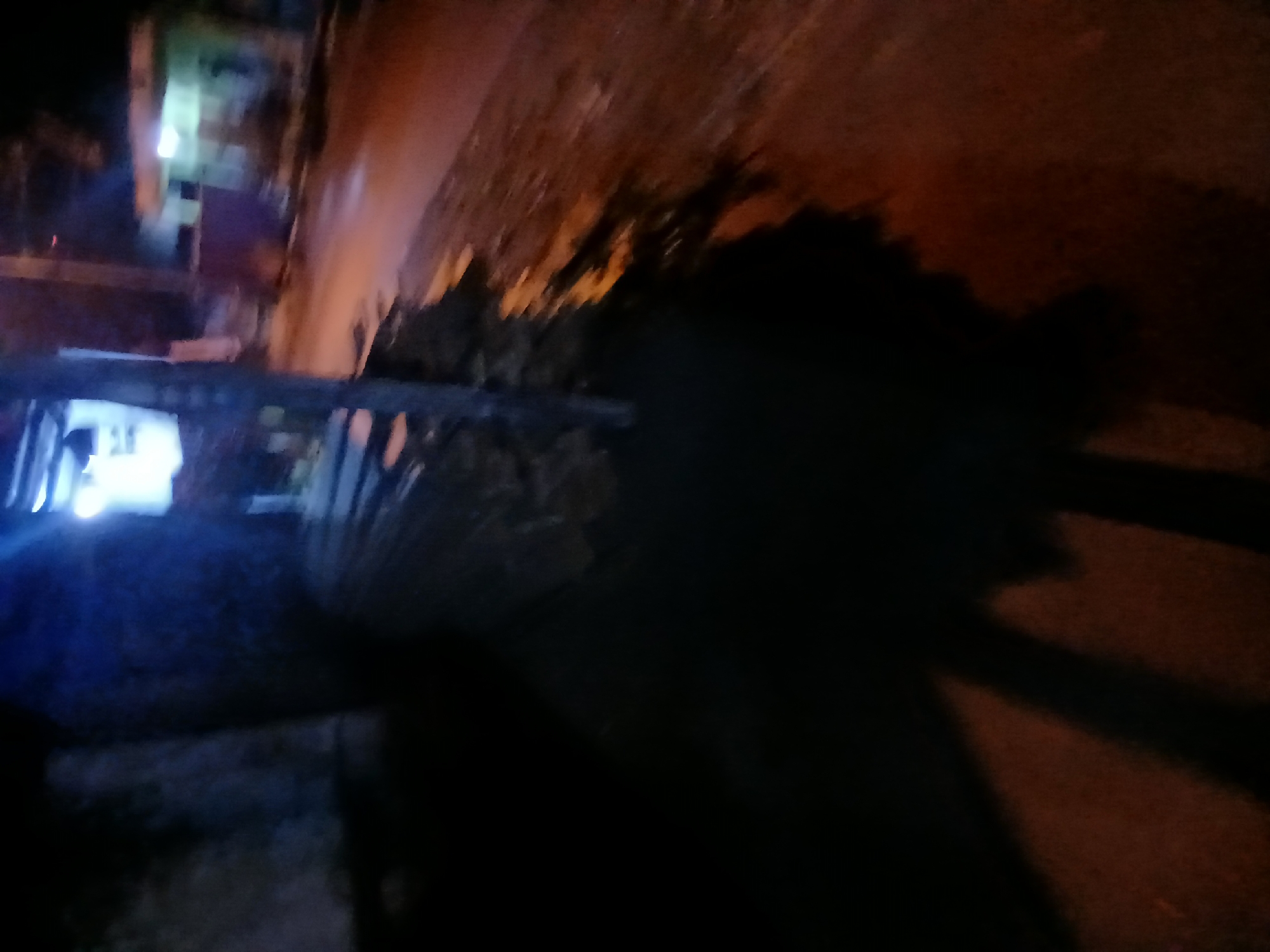 The image depicts a dimly lit, outdoor scene at night, possibly in Kademangan Kulon, Indonesia. In the foreground, a large, dark shadow covers much of the ground, suggesting the presence of an unseen object or person to the left. The shadow stretches across the reddish-brown ground, which appears to be a road or pathway. To the left of the shadow, a dark, textured surface is visible, possibly a wall or building.
In the midground, a stack of long, thin objects, likely wooden planks or similar material, is piled horizontally. These objects are dark and indistinct due to the low light. Behind the planks, a vertical pole or structure stands, illuminated by a bright, bluish light emanating from above and to the left.
Further in the background, a building with glowing windows can be seen. The light from the windows is white and illuminates the surrounding area. The overall impression is of a quiet, possibly residential or commercial area at night, with minimal ambient light. The blurriness of the image suggests it was taken with a hand-held camera in low light conditions.