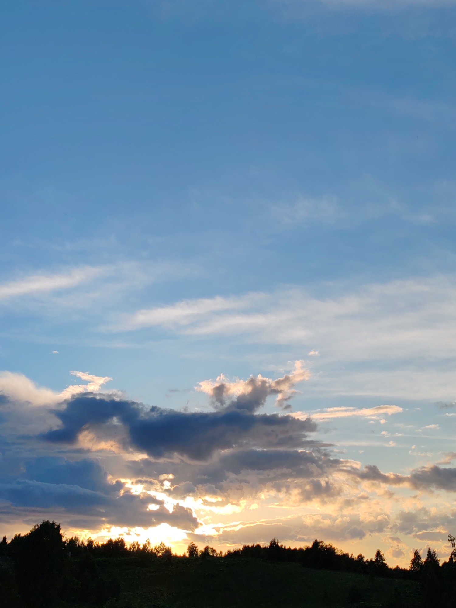 The image captures a sunset over a forested area in Chuchlema, Russia. The sky is a blend of pale blue and wispy white clouds, with streaks of orange and yellow light emanating from the setting sun peeking through darker, more substantial clouds near the horizon. The foreground is dominated by a silhouetted line of trees and dark green foliage, suggesting a hilly or uneven terrain. The overall impression is one of a serene natural landscape at dusk. There are no people or visible human activity in the frame.