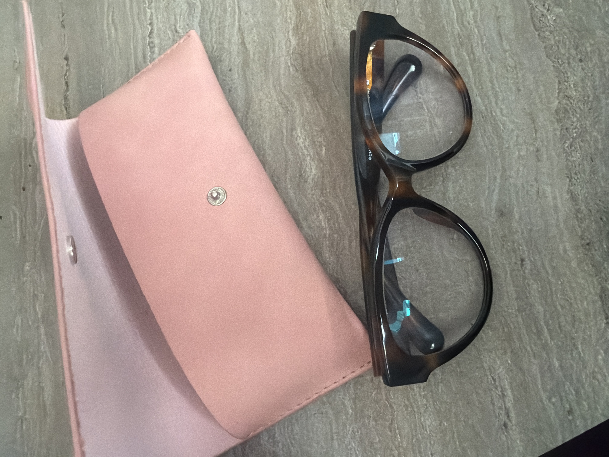 A pair of tortoiseshell-patterned eyeglasses with clear lenses and dark brown frames rests on a textured, beige and gray surface. The eyeglasses are positioned slightly angled, with the left lens closer to the viewer. A soft pink glasses case is open behind the eyeglasses, with its flap extended upwards. The case has a subtle sheen and appears to be made of a faux leather material. A small, silver snap closure is visible on the front of the case. The background surface has a stone-like appearance with visible marbling and slight indentations. There are no people present in the image, and no actions are being performed. The scene is static and focused on the objects.