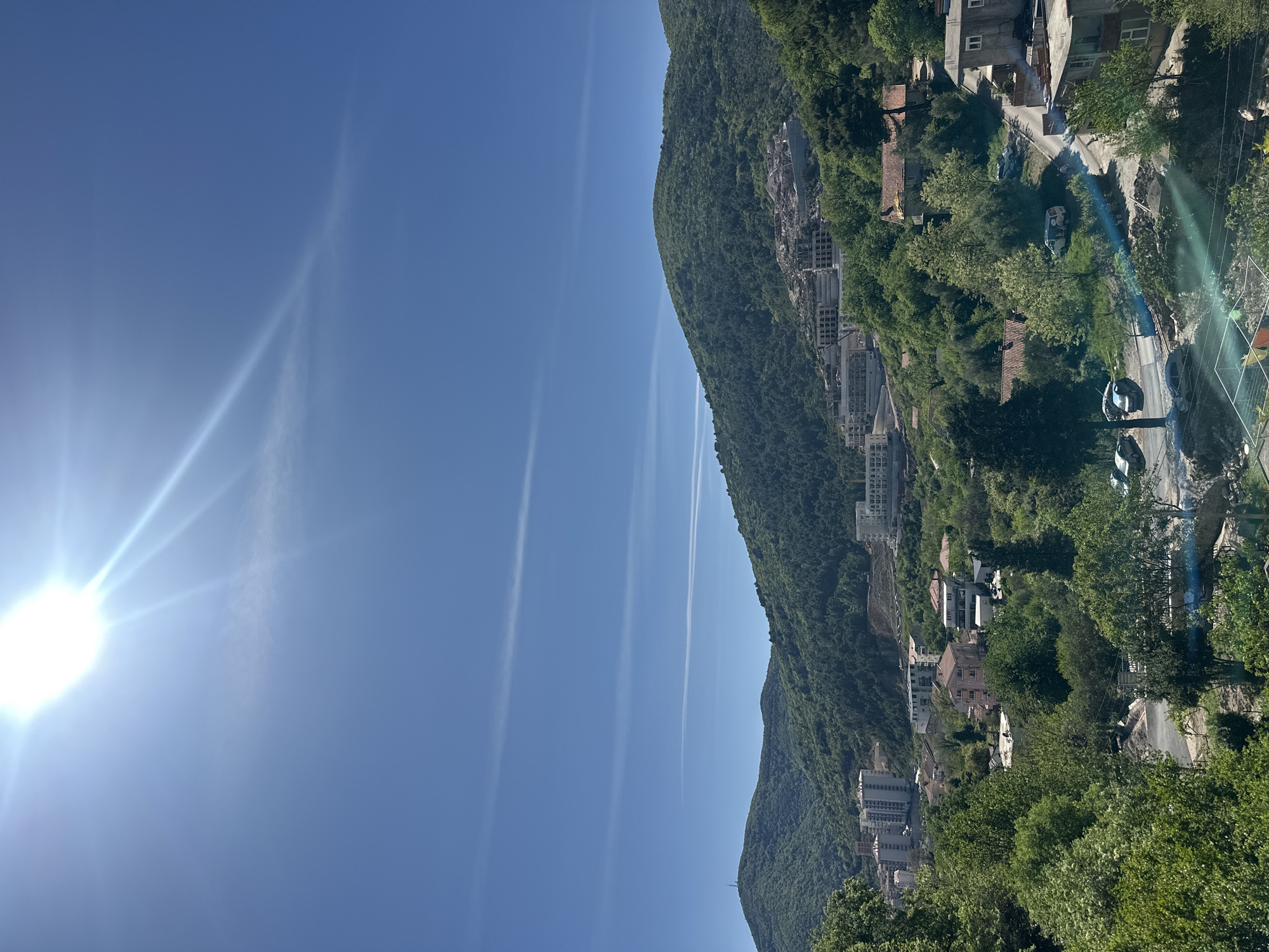 The image depicts a hillside view in Budva, Montenegro, on a clear, sunny day. The foreground shows dense green foliage and trees. A road winds through the foliage, with several cars parked along its edge. Mixed residential and commercial buildings are interspersed among the trees, indicating urban development along the hillside. A large, forested mountain rises on the right side of the frame. The sky is clear blue, with contrails from aircraft. The sun is visible in the upper left, emitting bright rays.
