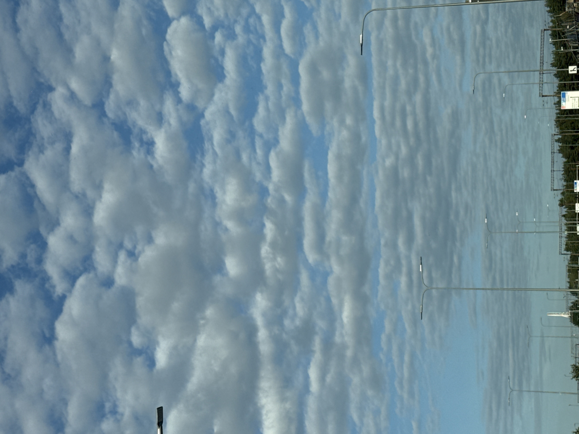 The image depicts a street scene in Tallinn, Estonia, on a cloudy day. A series of streetlights line the road, some of which are illuminated. The sky is filled with a pattern of cumulus clouds. In the background, there is a line of trees and some structures that appear to be industrial or transportation-related, such as power lines and possibly a train track. A white tower-like structure is visible in the distance. No people or vehicles are clearly discernible in the frame. The overall atmosphere is overcast, suggesting it is daytime but not necessarily bright.