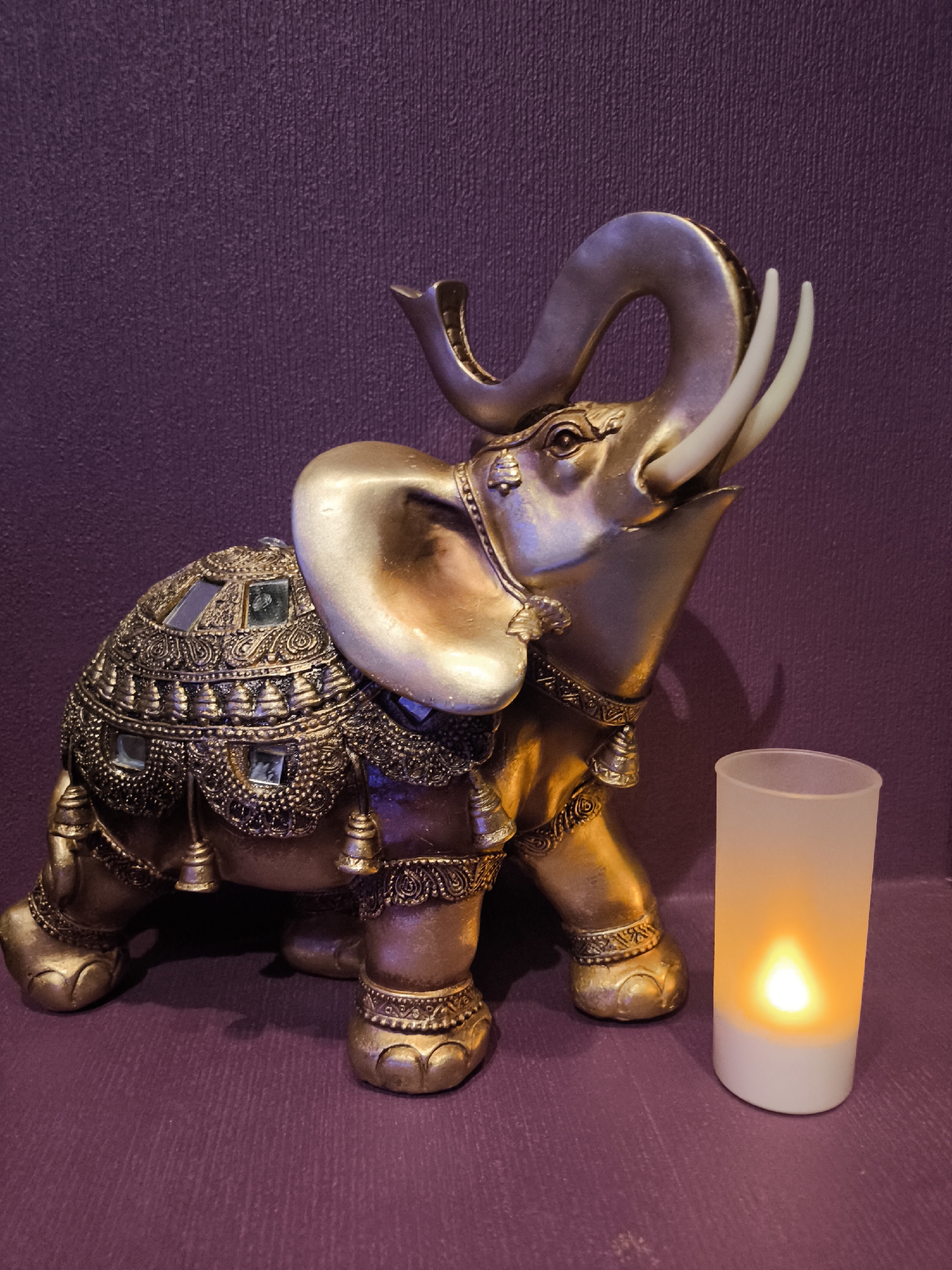 A gilded elephant statue stands next to a lit, frosted candle in front of a textured purple wall. The elephant has a decorative saddle adorned with embellishments and miniature bells. The location in Khimki, Russia, is not apparent from the image.