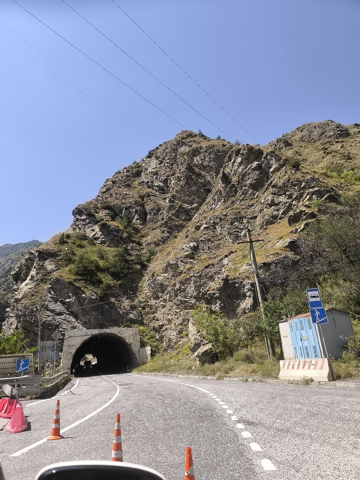 This image depicts a view of a mountain road leading to a tunnel entrance in Mizur, Russia. The road surface is asphalt with white lane markings. Several orange traffic cones are placed on the side of the road, indicating some form of traffic management or construction. A concrete barrier is also visible.
To the right of the road, there is a light blue electrical cabinet with some signage and a concrete block. Above the cabinet, a blue traffic sign shows a bus symbol and a pedestrian symbol with an arrow pointing to the right. A wooden utility pole with wires is also present.
The mountain face to the left and behind the tunnel is rocky and sparsely vegetated with dry grass and some green bushes. Power lines are strung across the sky. The sky is clear and blue. A faint crescent moon is visible in the sky towards the upper right.
The tunnel entrance is a concrete archway. Inside the tunnel, the headlights of a vehicle are visible, indicating that traffic is proceeding through it. The overall scene suggests a remote, mountainous area with active road infrastructure.