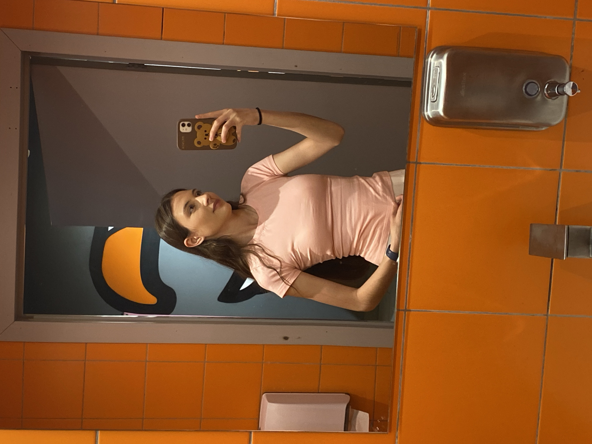 A woman is taking a selfie in a bathroom mirror. The bathroom has orange tiled walls. A stainless steel soap dispenser is on the wall. The mirror reflects a gray wall with a large orange and black graphic of a figure reminiscent of the video game "Among Us". The exact location in Votkinsk, Russia is not ascertainable from the image.