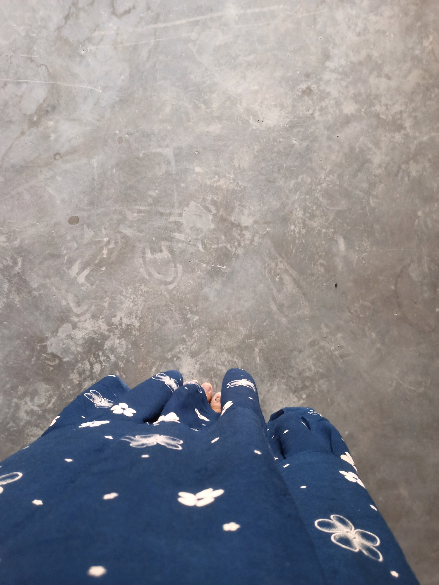 A high-angle, outdoor shot shows the lower half of a person's legs and feet wearing a dark blue skirt with a white floral pattern. The skirt is gathered and bunched around the legs. The ground beneath is a textured concrete surface with visible marks and some small debris. The scene appears to be located in Karangtalun, Indonesia.