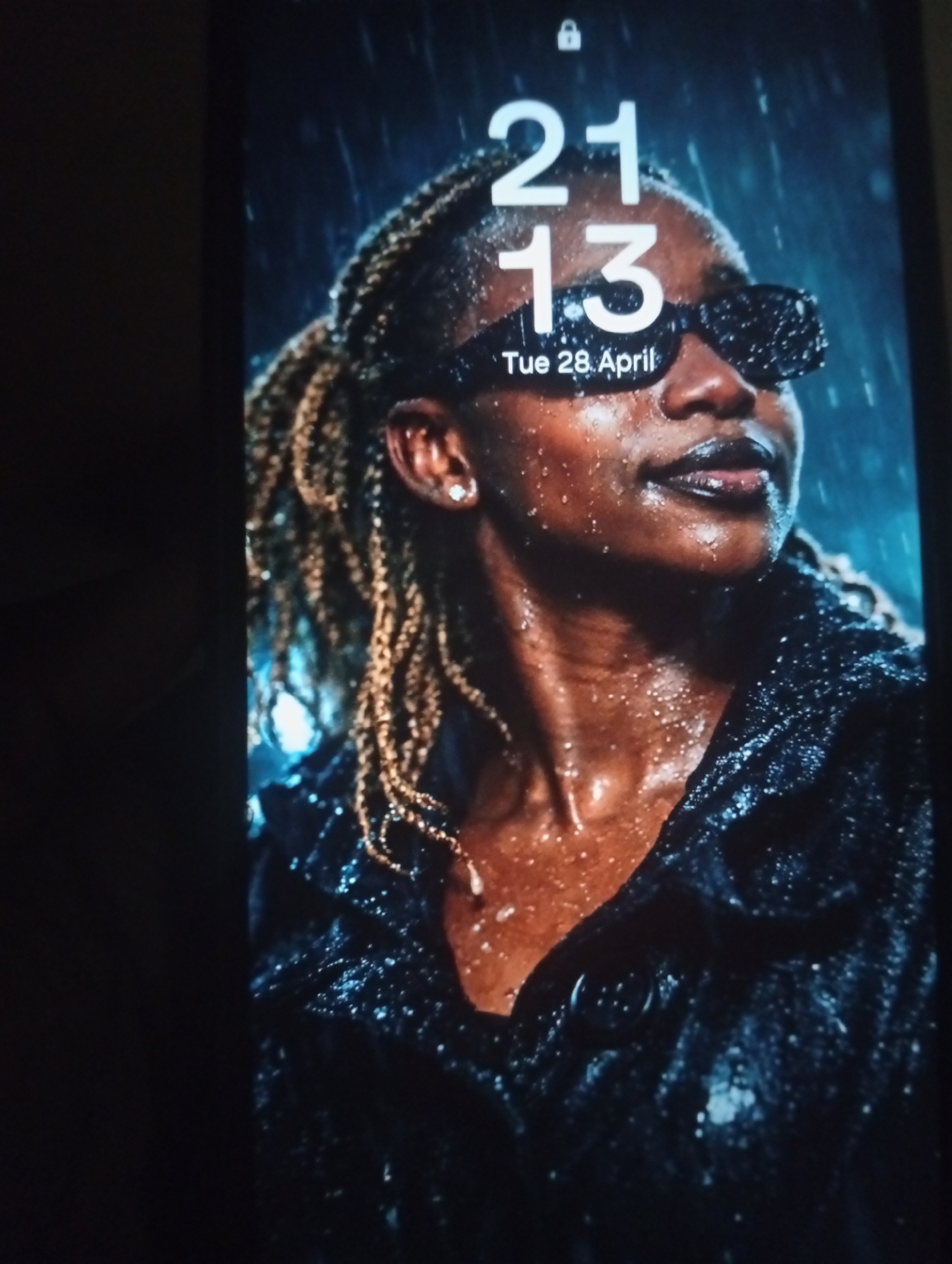 The image displays a smartphone screen showing a lock screen wallpaper featuring a close-up portrait of a person, likely a woman, under what appears to be rain. The lock screen text indicates the time "21 13" (9:13 PM) and the date "Tue 28 April" in white font, with a small white padlock icon at the top center.
The subject has dark skin and is looking upwards and slightly to the right with a focused or pensive expression. She wears dark, rectangular sunglasses that obscure her eyes, and her lips appear to have a dark, glossy finish. Her hair is styled in numerous small, light brown or blonde braids or locs that fall around her face and shoulders. A small, round stud earring is visible in her right ear.
Numerous water droplets are evident on her skin, particularly on her forehead, cheeks, neck, and shoulders, glistening as they catch the light. Her dark clothing, possibly a jacket or coat with a textured or sequined surface, also appears wet and covered in droplets. The background is dark and out of focus, suggesting a low-light environment, with subtle hints of blue and green light. The overall scene conveys a dramatic and stylized mood, likely a professional photograph or still from a video.