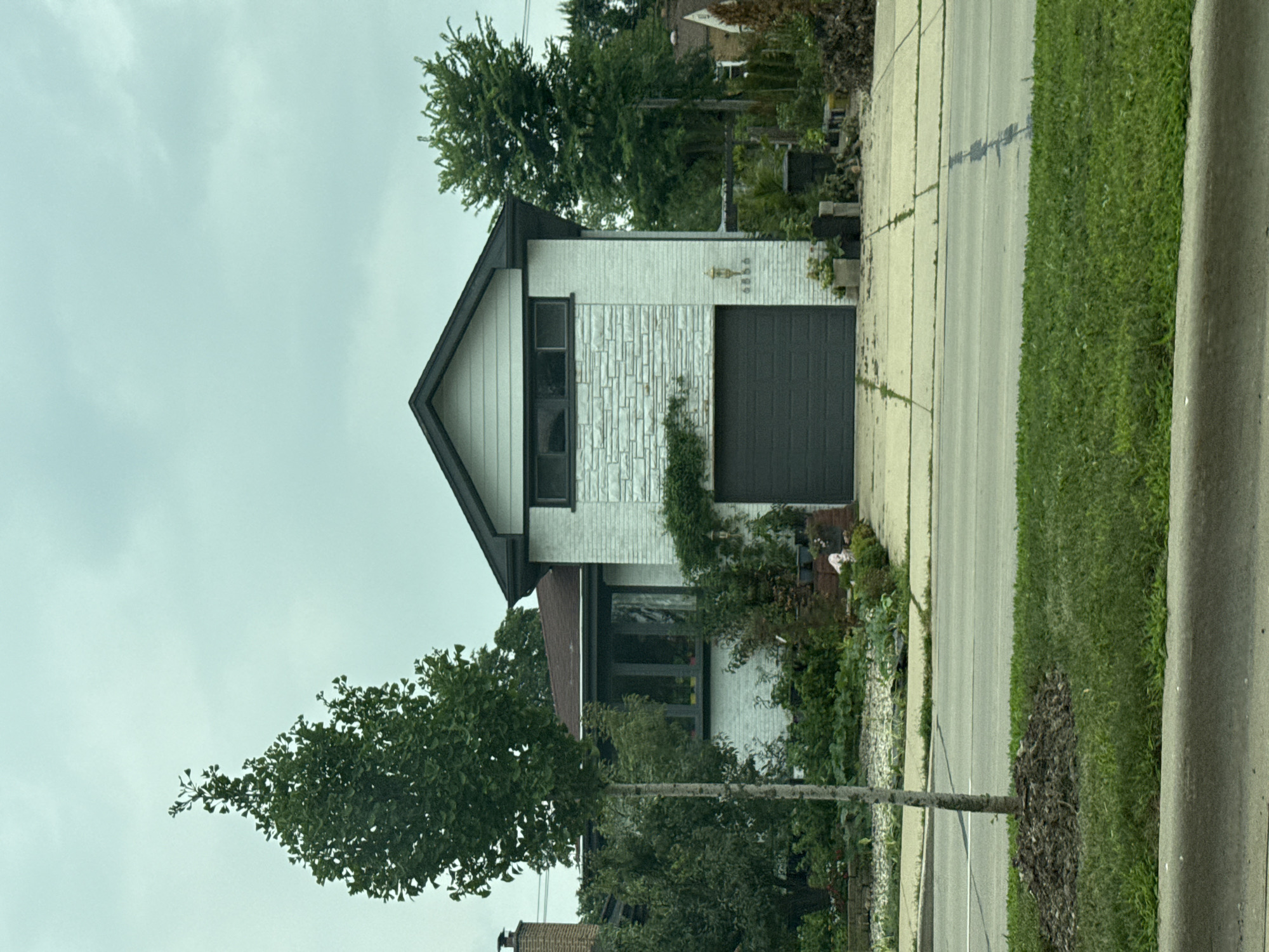This is a full, eye-level, vertical, outdoor, daytime, medium shot of a modern, two-story house with a garage in Lincolnwood, Illinois, USA. The house has a white brick facade on the second story and a light grey stone facade on the first story. A dark grey garage door is centrally located on the first story. A small, young tree with a thin trunk stands in the foreground to the left of the house. Lush green grass covers the area in front of the house and a sidewalk runs in front of the garage. The sky is overcast and grey, suggesting cloudy weather.