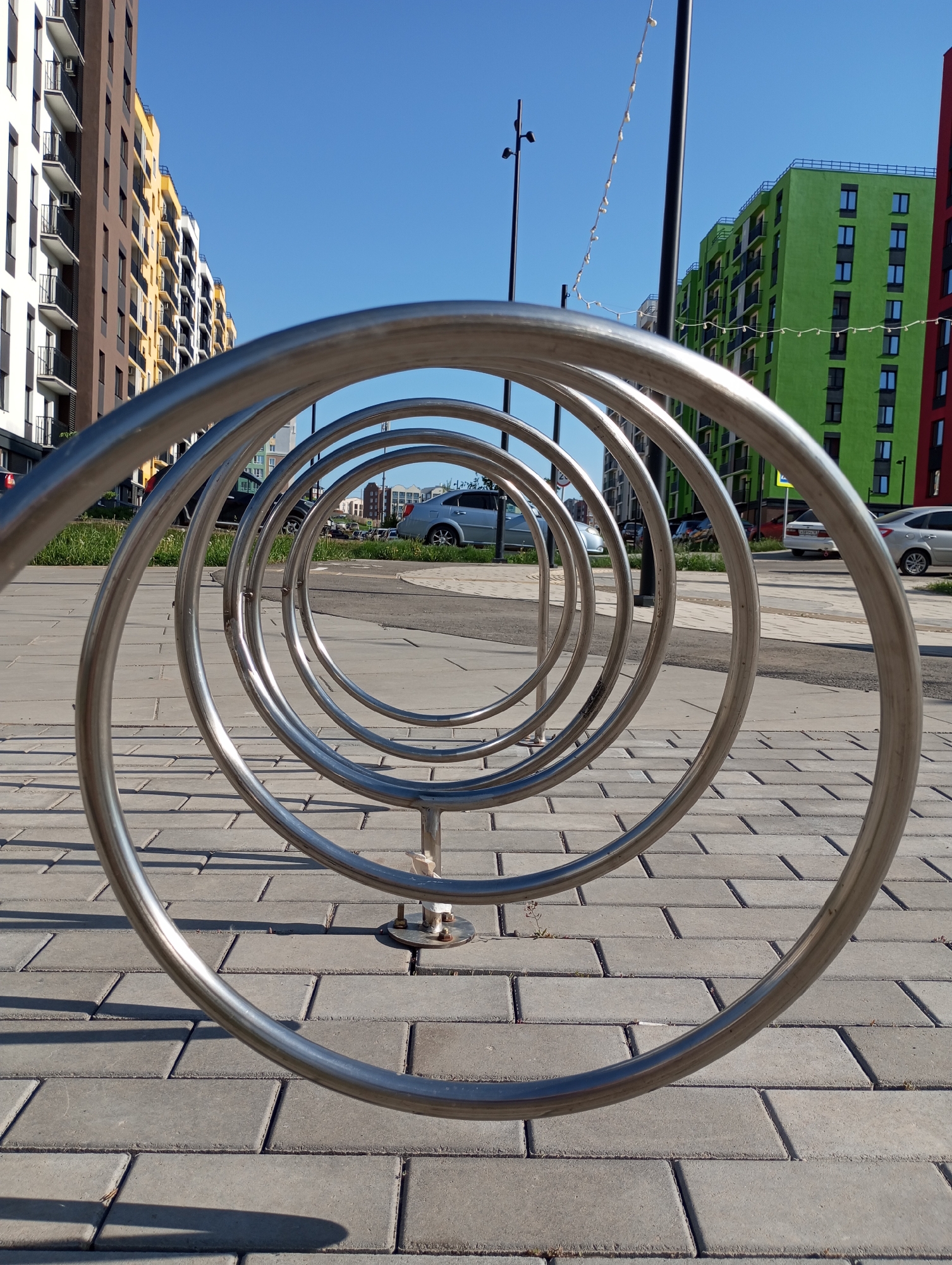 A bicycle rack consisting of five nested metal rings stands on a paved area in Pervomaysky, Russia. The pavement is made of gray rectangular blocks. Behind the rack are residential buildings with varying colors, including brown, white, yellow, green, and red. Cars are parked on the road behind the bike rack and buildings. Streetlights are visible in the background. The sky is clear and blue.
