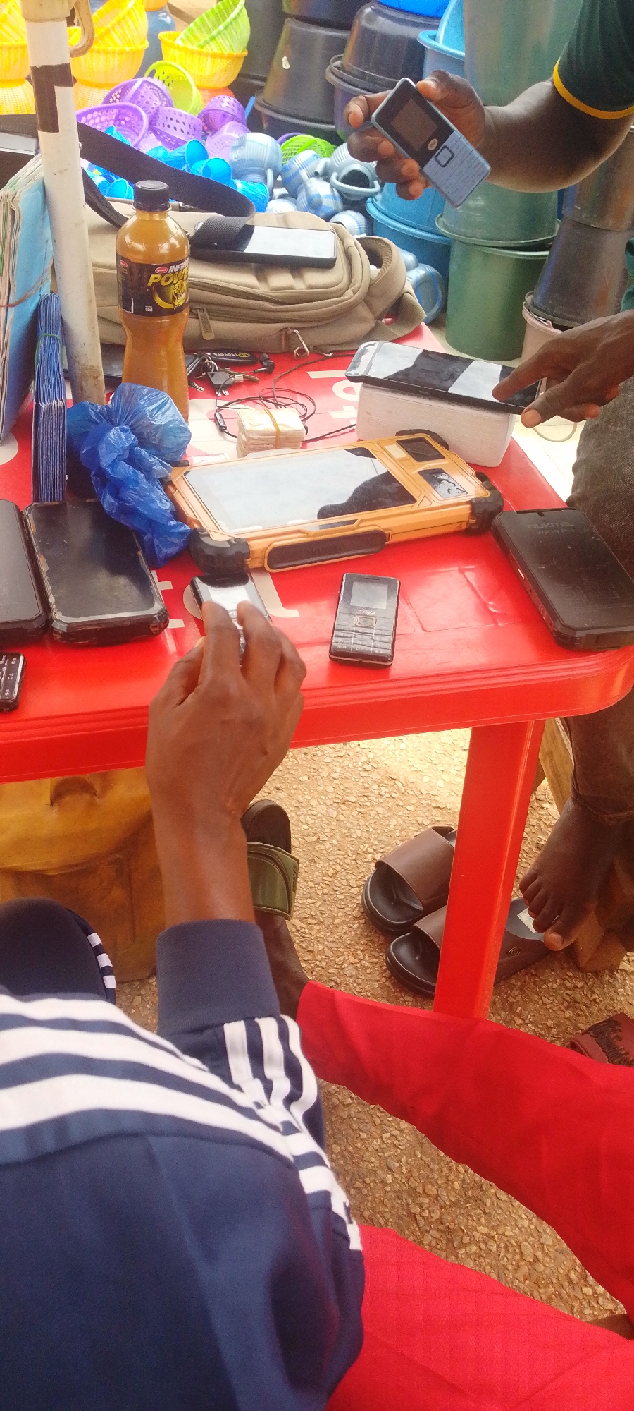The image depicts a transaction taking place at an outdoor market stall, likely in Baissa, Nigeria. The primary subjects are several individuals, their hands and forearms visible as they interact with various electronic devices and goods. A red plastic table dominates the foreground, laden with a collection of cell phones, a tablet device in a rugged yellow case, a bottle of "Infinity Power" drink, keys, and some bundled currency notes. In the background, a variety of plastic containers, baskets, and buckets are stacked, suggesting a stall selling household items or supplies.
The activity centers around what appears to be a mobile money transaction or a sale of electronic devices. Hands are seen holding and operating cell phones, with one person seemingly about to use a phone on the table. The presence of currency and the diverse electronic devices hints at commerce.
Notable details include the bright lighting, suggesting it is daytime. The ground is covered with gravel, indicative of an outdoor setting. The subjects are dressed in casual attire, with one person wearing a blue and white striped shirt and red trousers. The overall scene conveys a sense of a busy market environment focused on technology and daily commerce. The visible text on the drink bottle reads "INFINITY POWER".
