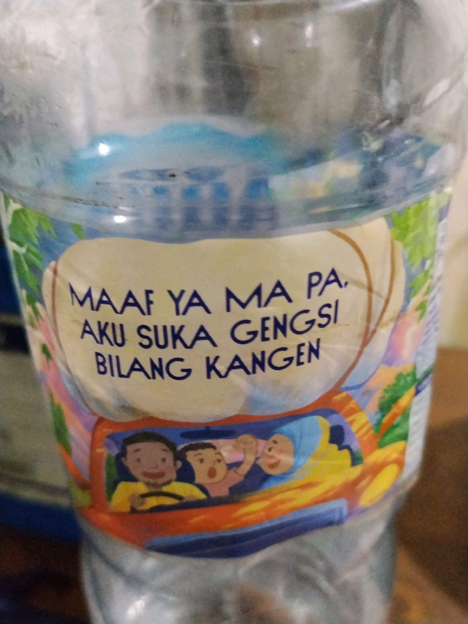 The image shows a close-up of a plastic bottle with a label. The label features a cartoon illustration of people in a car and text written in Indonesian. The text reads "MAAF YA MA PA, AKU SUKA GENGSI BILANG KANGEN," which translates to "Sorry Mom and Dad, I like to pretend I don't miss you." The bottle appears to be transparent and contains a clear liquid. The background is blurred, but hints of a wooden surface and possibly other objects suggest an indoor setting. The location of Sempayau, Indonesia, is not visually represented in the image itself; it is an external piece of information.