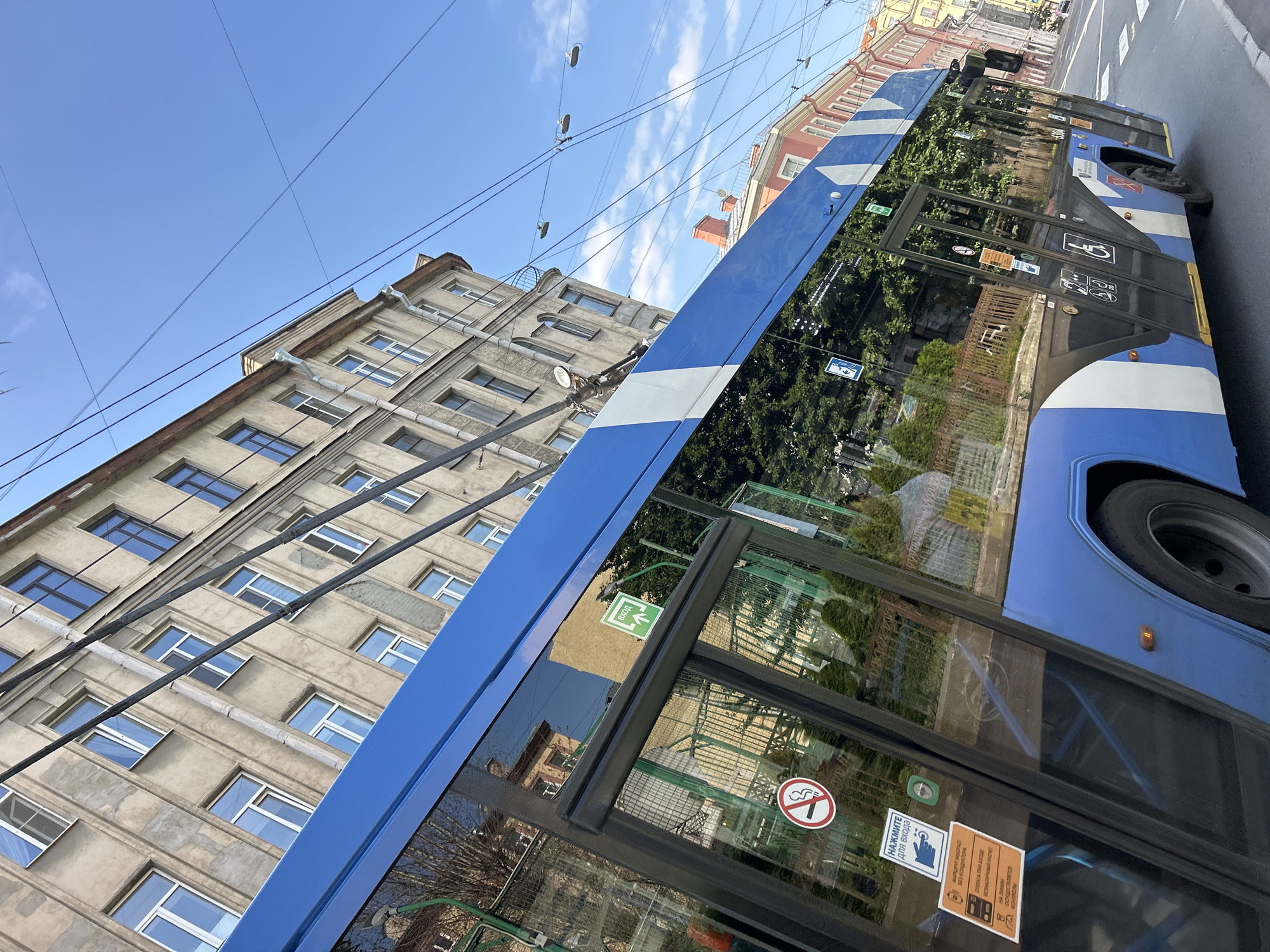 The image shows a blue and white bus on a city street. The bus is seen from a low angle, with its side facing the viewer. The windows of the bus reflect the surrounding buildings and trees, indicating a sunny day. The bus is equipped with overhead wires, suggesting it is an electric trolleybus.
On the side of the bus, several signs are visible. One sign has an arrow pointing downwards with the word "Вход" (Entrance) written above it in Cyrillic. Another sign shows a smoking symbol crossed out, indicating "No Smoking". Below the entrance sign, there is another sign with text in Cyrillic that reads "НАЖМИТЕ для входа" (Press to enter). There are also signs related to fare payment and route operation.
The bus is parked next to a tall, old building with many windows. The building appears to be residential or office space, with a weathered facade. Overhead, there are numerous electric wires, further confirming the presence of a trolleybus system. The sky is blue with some scattered white clouds. The overall impression is that of an urban environment, likely in Eastern Europe, given the Cyrillic script. The image appears to have been taken during the daytime, likely in the morning or afternoon, judging by the sunlight. There are no people visible in the frame, and the bus appears to be stationary.
---
*Note: The image has been rotated 90 degrees clockwise.*