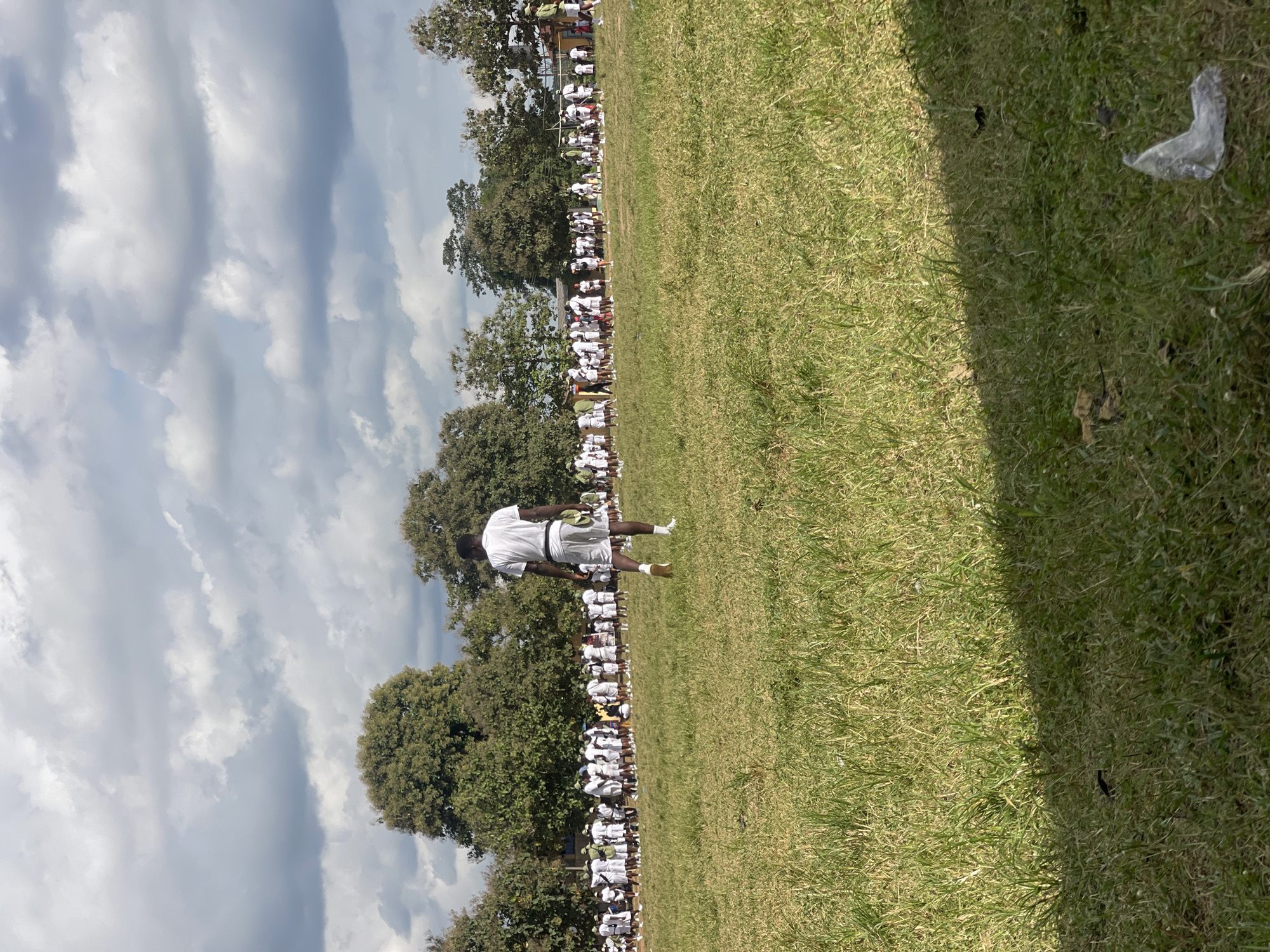 In Emure-Ekiti, Nigeria, a group of people, likely young adults, are assembled in a large open field. The ground is covered in green grass, with some patches appearing dry and yellowed. The individuals are predominantly dressed in white shirts and white shorts or skirts, suggesting they may be participants in an event or training program. They are standing in rows, forming lines that stretch across the field. In the foreground, a single individual, also in white attire, is walking away from the camera, facing the assembled group. Trees with dense green foliage are visible in the background, and a cloudy sky with patches of blue dominates the upper portion of the image. The scene appears to be outdoors, possibly during the daytime.