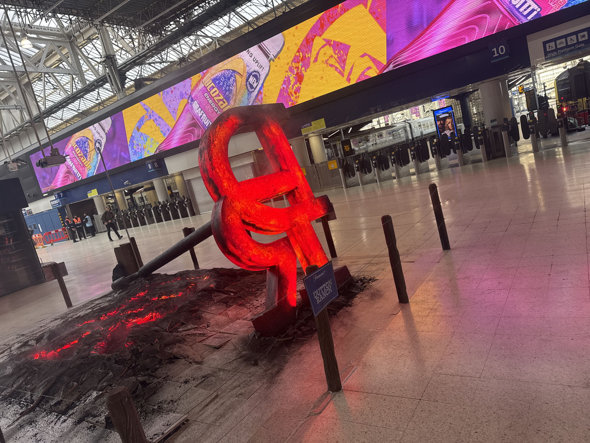 The image shows an indoor scene at a train station, likely in the city of London, United Kingdom. In the foreground, a large, red, stylized sculpture of the ampersand symbol (&) is surrounded by dark, irregular material that mimics molten lava, with red lights illuminating it to enhance the effect. A small sign labeled "DUTTON RANCH" is placed nearby, likely a promotional display for a television show.
In the background, there are several rows of ticket barriers leading to the platforms. A large digital advertising screen displays promotional images for the Lucozade drink. The architectural structure of the train station, with its high, metal-framed roof, is visible in the upper part of the image. The floor is tiled, and there are several dark wooden posts or barriers surrounding the sculpture. People can be seen in the distance walking toward the platforms. Signage indicating platform numbers and information is also visible. A train sits at one of the platforms.