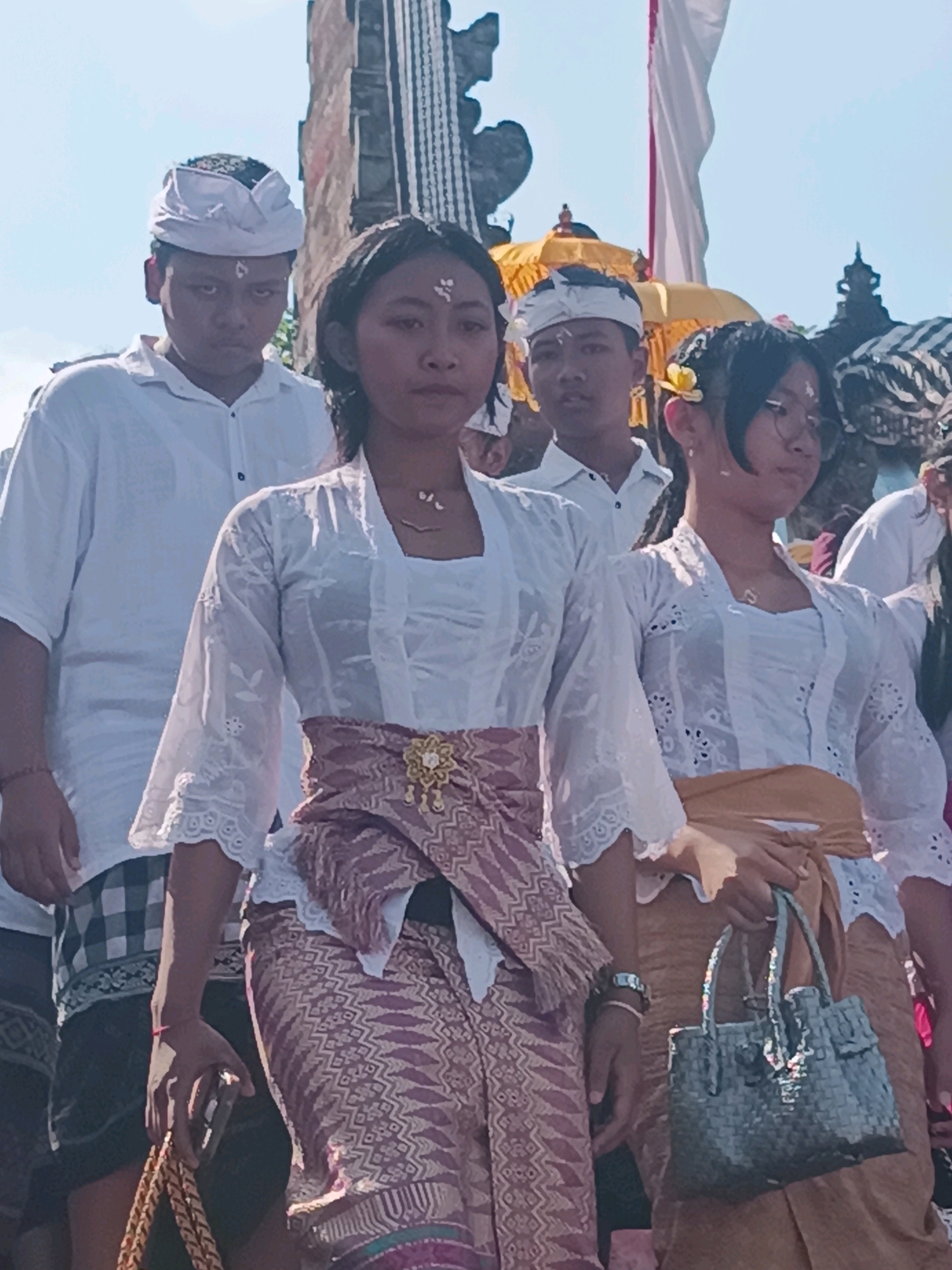 The image depicts a procession or ceremony at a temple in Tabanan, Indonesia, likely on a sunny day. Several young people, dressed in traditional Balinese attire, are featured. The foreground is occupied by two young women and a young man. The woman in the center wears a white lace kebaya and a patterned sarong, cinched with a decorative belt. To her right, another young woman, also in a white kebaya and a golden sarong, carries a woven grey handbag. Behind them, a young man wears a white shirt and a white headcloth (udeng). Further back, another young man in similar attire is visible. The background includes elements of temple architecture, such as stone carvings and a white banner, suggesting a religious or cultural event. The overall atmosphere appears to be one of solemnity and tradition.