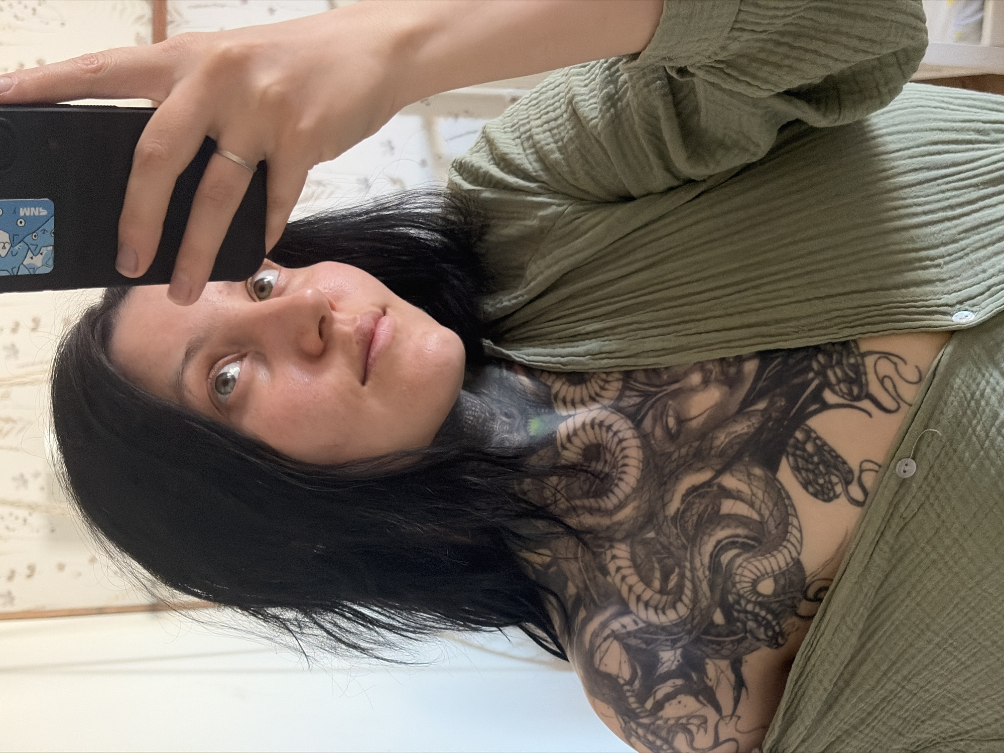 A woman with dark hair and a large tattoo of snakes covering her neck and upper chest is taking a selfie. She is holding a black smartphone in her left hand, with a sticker of cartoon figures on the back. She is wearing a sage green, textured button-up shirt. The background appears to be a light-colored wall with faint patterns. The image does not provide information confirming the location is Chaikovsky, Russia.