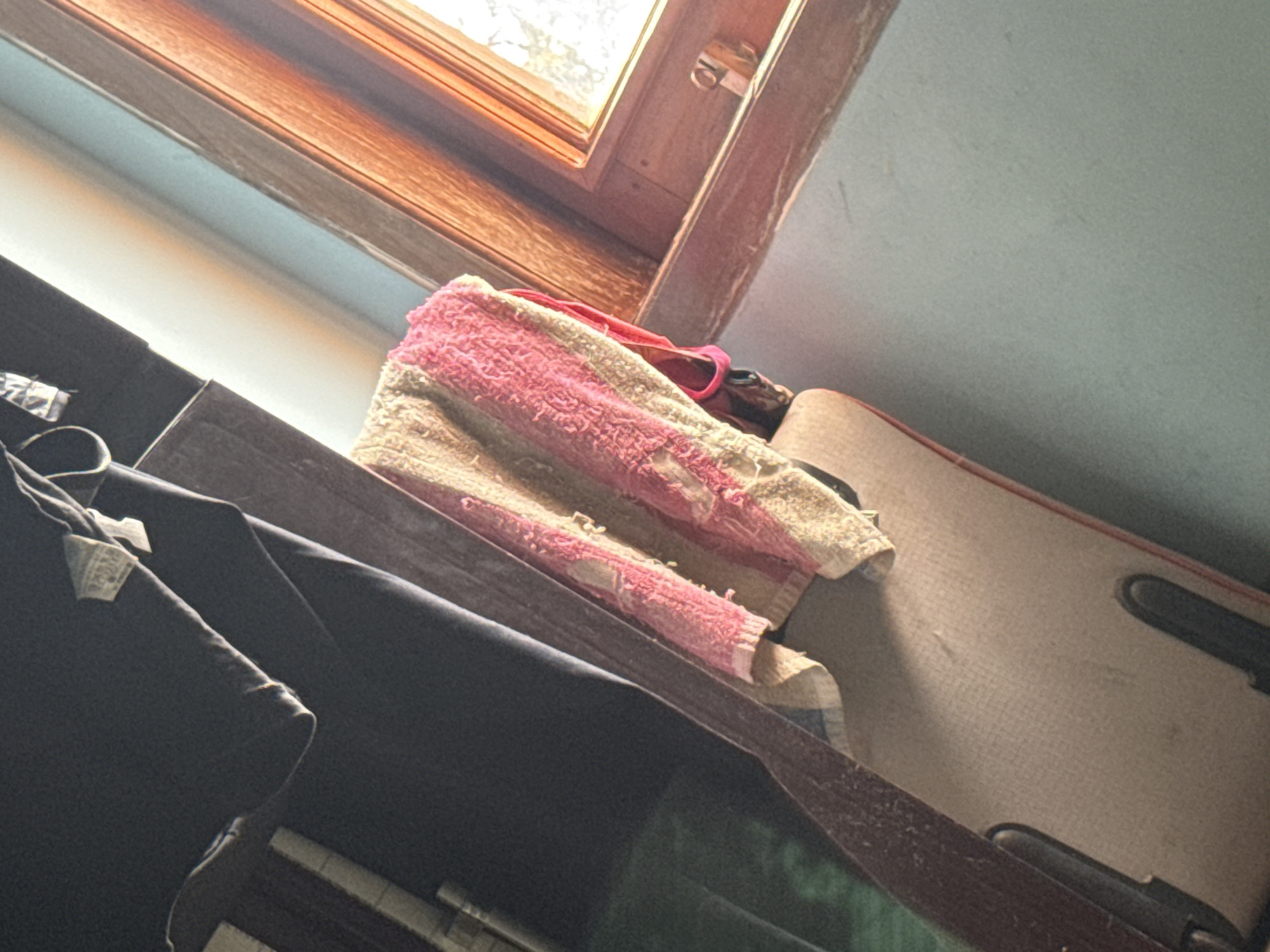 The image captures an indoor scene, possibly a room in Slahung, Indonesia, characterized by warm lighting filtering in from a window. In the foreground, a textured towel with pink and beige stripes is draped over the back of an upholstered chair. The chair itself appears to be made of wood and fabric, with dark accents. Behind the chair, a dark, draped piece of clothing hangs, its texture suggesting fabric. The background is dominated by a light blue wall and a wooden window frame with sunlight streaming through. The overall impression is one of domesticity, with the objects suggesting a lived-in space. The perspective is slightly tilted, giving the image a dynamic feel.