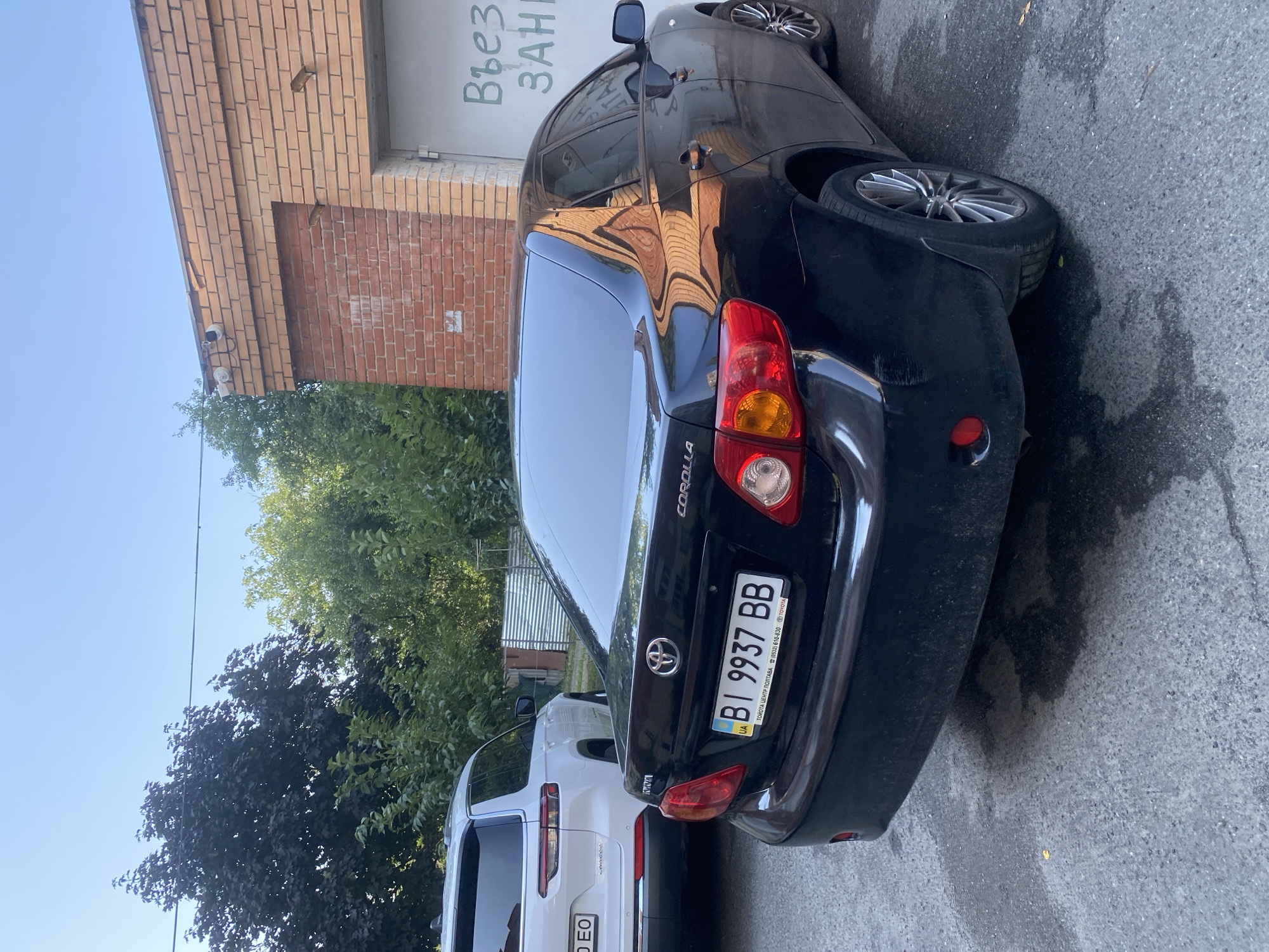 The image shows the rear of a black Toyota Corolla parked on an asphalt surface, likely in a city. The car's license plate is visible and reads "BI 9937 BB". Next to the Corolla, the rear of a white SUV is visible, with a license plate displaying "0 EO". In the background, a brick building with a garage door is present. Above the garage door, two security cameras are mounted. Green trees and a clear sky are also visible in the scene. The overall environment appears to be an outdoor urban setting. The building in the background has Cyrillic lettering written on its garage door. The scene appears to be set in Poltava, Ukraine, based on the license plate formatting which is consistent with Ukrainian plates.