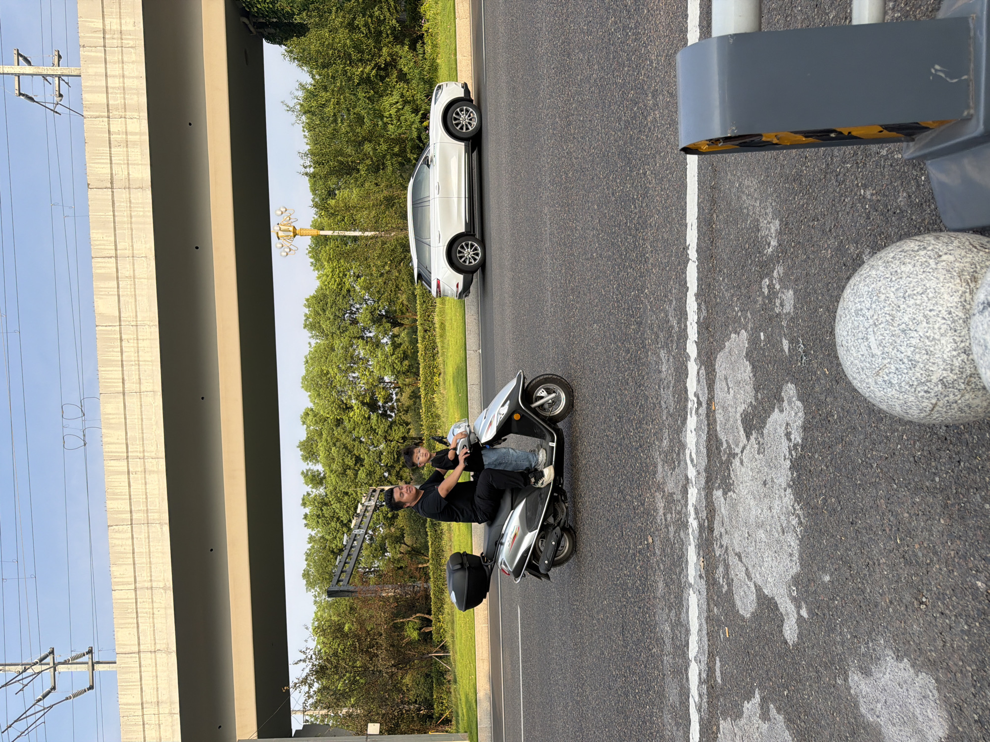 In Jiangjin, China, under an elevated overpass with overhead electrical lines, a man and a young boy ride a silver scooter on a paved road. The man, wearing a black t-shirt and pants, drives the scooter, while the boy, in a black t-shirt and jeans, sits behind him. Both are wearing masks. A white SUV is visible driving in the background. Lush green trees line the roadside, and the sky is clear blue. In the foreground, a portion of a decorative boulder and a gray traffic barrier with yellow and black hazard stripes are visible.