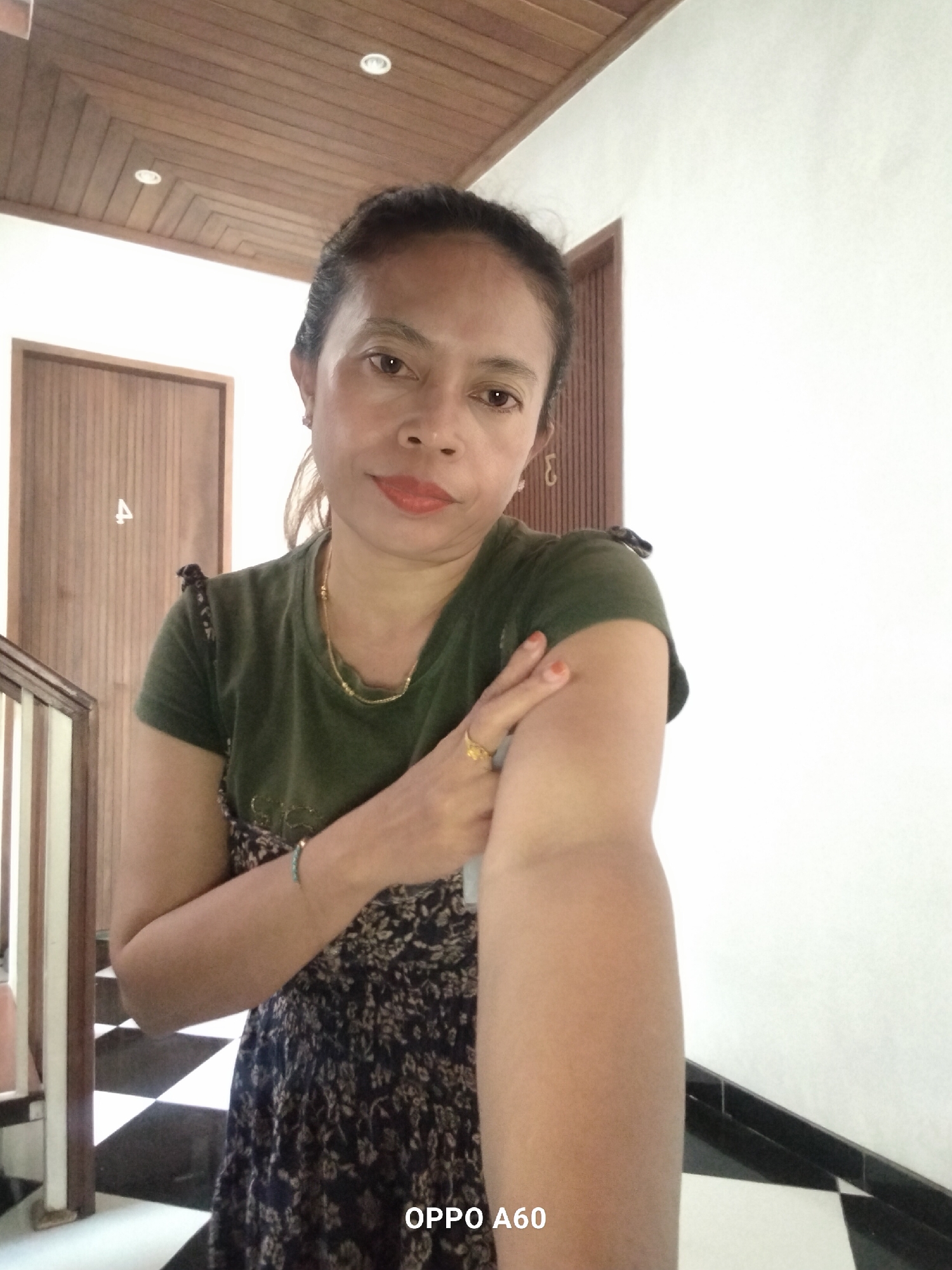 A medium shot photograph shows a woman standing indoors, facing the camera. She has dark hair pulled back, light brown eyes, and is wearing a dark green t-shirt with a floral print dress underneath. She is wearing a gold necklace, a gold ring on her right hand, and a blue bracelet on her left wrist. The woman is looking directly at the camera with a neutral expression. She is holding her right arm with her left hand, her left arm is bent at the elbow.
The background consists of a wooden ceiling with recessed lighting, a white wall, and a wooden door. To the left of the woman is a staircase with wooden banisters and a checkered pattern on the steps. The image is lit with natural light. The woman's arm is positioned to her right, towards the camera. The lower right corner of the image displays text that reads "OPPO A60". The context suggests this photo was taken in North Kuta, Indonesia.
