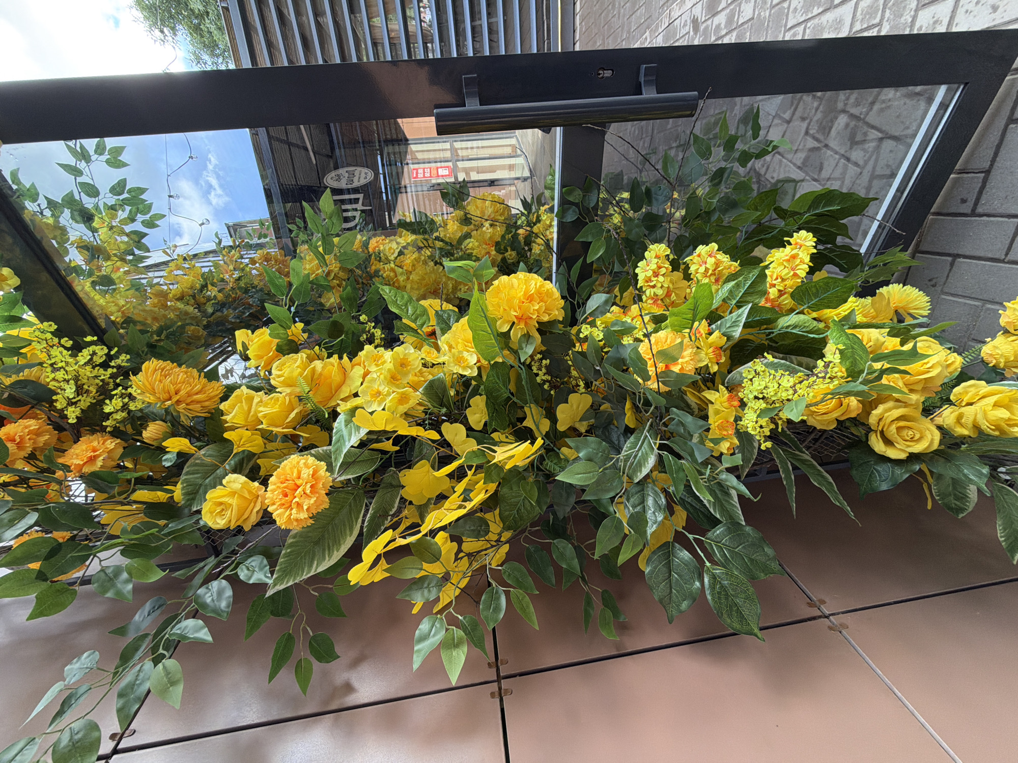 A vibrant floral display of artificial yellow and orange flowers with abundant green foliage is captured in bright daylight. The arrangement is situated in front of a dark, rectangular structure, which features a highly reflective surface. This surface acts as a mirror, reflecting a clear blue sky with scattered white clouds, a slatted overhead structure, and a patterned brick wall, suggesting an outdoor or semi-outdoor setting like a building facade or patio.
The flowers include various types of yellow blossoms, such as roses, chrysanthemums, and clusters of smaller, bright yellow and orange blooms, complemented by broad and trailing green leaves. On the right side of the black frame, a horizontal bar or handle is visible, along with a small light or sensor. The ground beneath the display is made of light brown or beige tiles, with distinct grout lines and small connectors visible.
In the reflections on the left side of the frame, a white circular sticker or logo is partially visible, containing text that appears to include "PUPURI" and "JAPAN." Deeper in the reflection, a red sign with the prominent Cyrillic letters "ТЭЦ" is discernible, indicating a location associated with a thermal power plant. The scene is devoid of people or activity, focusing solely on the decorative floral arrangement.
