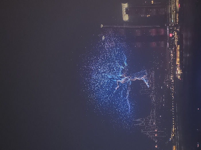 The scene is captured at night, depicting a large-scale aerial drone light show over a city located within 江北城街道, China. The dark sky is prominently illuminated by two distinct drone formations. The central formation presents a large, stylized tree composed of numerous small lights, with its extensive canopy rendered in blue and its trunk and main branches in orange and reddish hues. To the left of the tree, a second drone formation outlines a cityscape silhouette, depicting multiple illuminated high-rise structures and a bridge, also composed of similar small lights.
Below these aerial displays, a densely illuminated urban landscape stretches across what appears to be a wide river. The city skyline features numerous high-rise buildings extensively lit with a variety of white, yellow, and red architectural lights. On the far right, a tall, modern building with prominent horizontal yellow-gold light bands near its upper section is visible. Several large, illuminated vessels are present on the surface of the water. A tall, slender light pole or streetlamp with a curved top is visible on the right side of the image, rising from the mid-ground cityscape. No individuals are discernible within the frame.