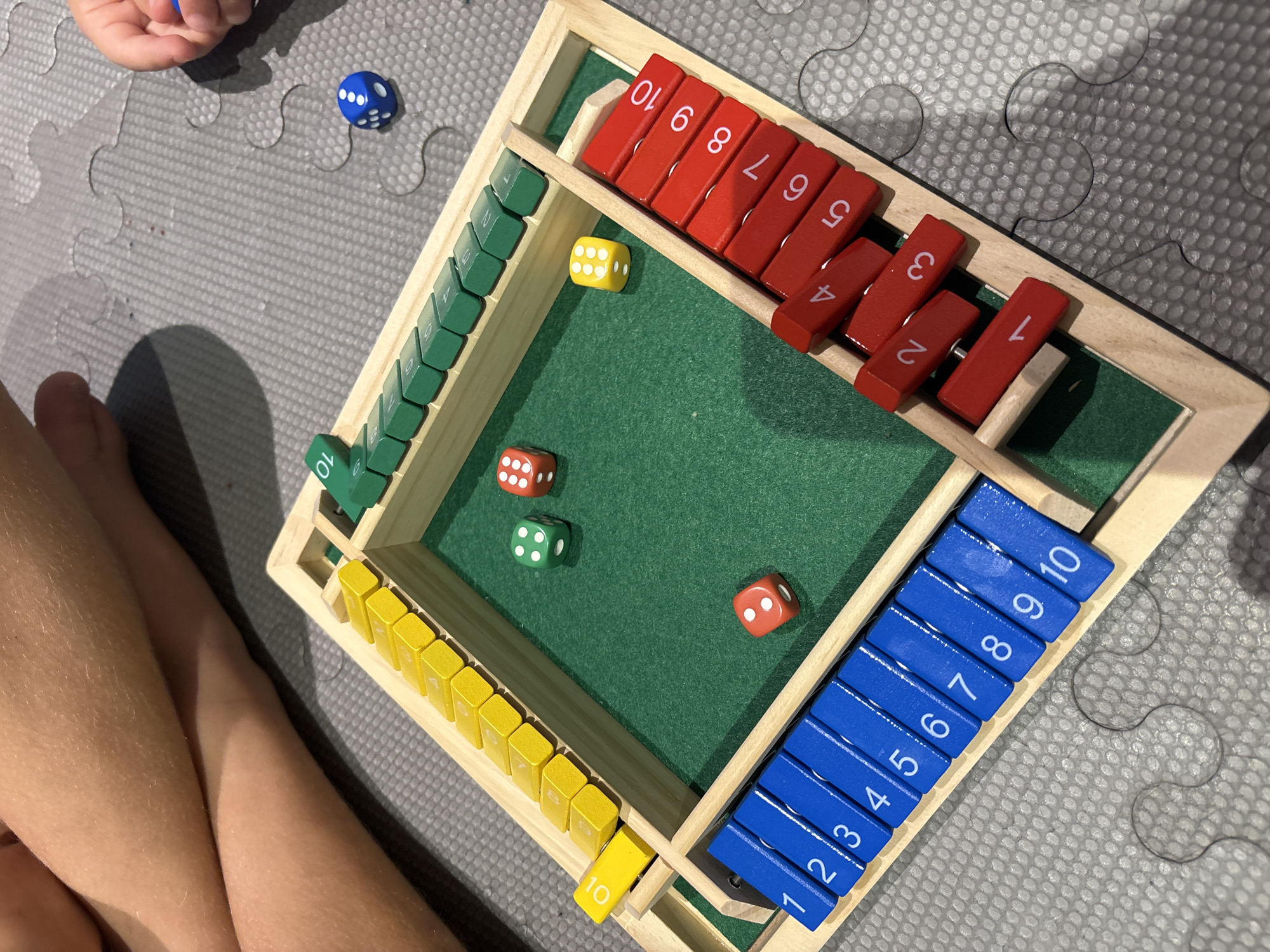The image displays a close-up, overhead view of a children's dice and scoring game on a gray puzzle mat. The game board is made of wood with a green felt playing surface. On the playing surface, there are three dice: one green with 4 dots, one red with 3 dots, and one red with 5 dots. Around the perimeter of the board are wooden tiles numbered 1 through 10, arranged in sections of different colors. There are yellow tiles on the top left, green tiles on the top right, blue tiles on the bottom left, and red tiles on the bottom right. The blue tiles are numbered 1 through 10. The red tiles are numbered 1 through 10, with only tiles 1 through 10 visible. A yellow tile labeled "10" is placed at the end of the yellow section. A blue die with 3 dots is visible on the gray mat to the right of the game board. In the background, the legs of two children are visible, suggesting they are playing the game. The environment is indoors, and the lighting suggests it could be daytime. The image does not provide any information about the location in Dubai, United Arab Emirates.