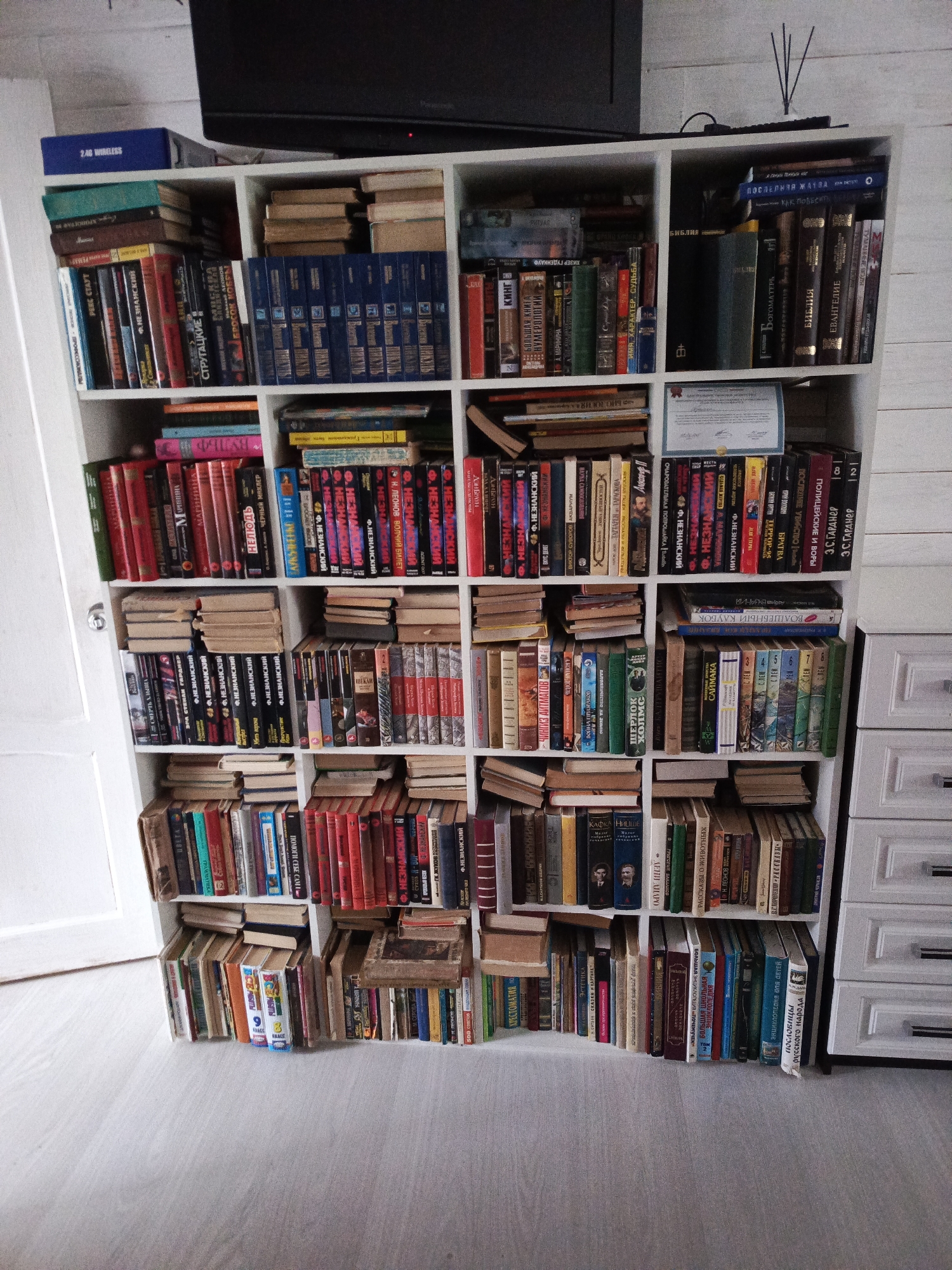 A floor-level, full shot shows a white bookcase filled with books, possibly in a residence. The bookcase has a grid-like structure, with shelves and compartments packed with books.
The books vary in color, size, and thickness, suggesting a diverse collection. Many books are stacked horizontally atop upright books. Some books have their spines facing the viewer, revealing titles and author names in Cyrillic script.
A flat-screen television sits on top of the bookcase.
The bookcase is positioned against a wall with white horizontal panels, and to the left of a white door with a silver handle. To the right is a white dresser.
The floor is covered with light gray flooring.
The location may be in Северный, Russia, as indicated by the OCR content in the image.