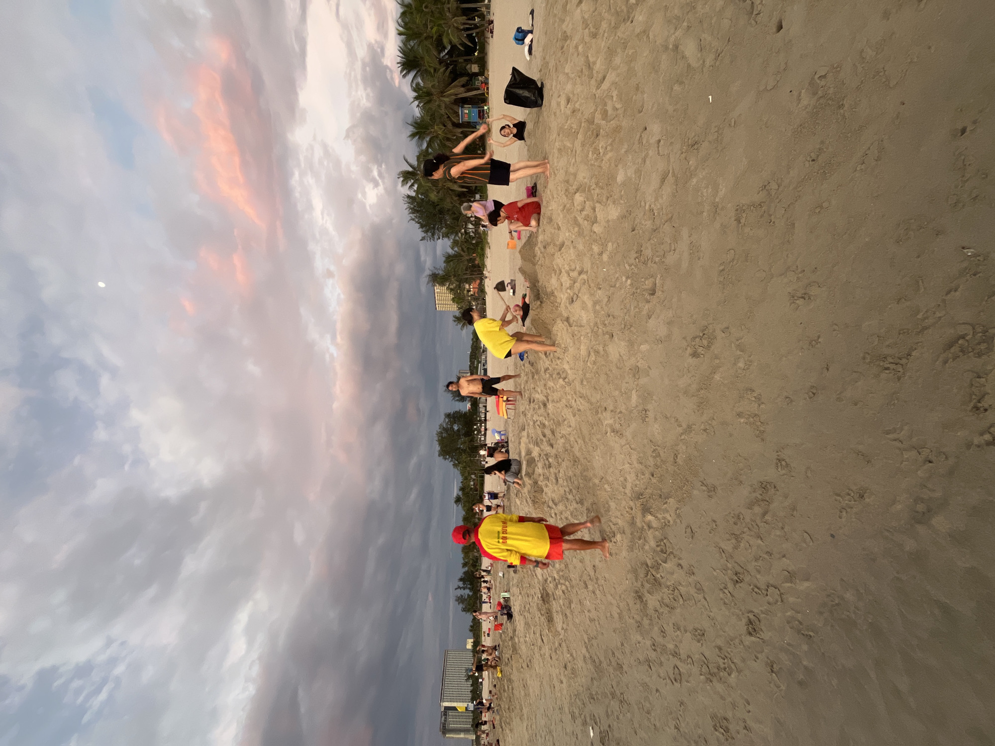 This image captures a vibrant beach scene in Đà Nẵng, Vietnam, during what appears to be either sunset or sunrise, indicated by the soft, warm light and streaks of orange and pink in the clouds. The sky is partly cloudy, with varying shades of grey, white, and a pale blue peeking through, and a faint moon is visible in the upper left.
The foreground is dominated by a wide expanse of light brown sand, showing subtle textures and footprints. Prominently featured in the lower center is a person facing away from the camera, wearing a bright yellow long-sleeved shirt with red sleeves and a red peaked hat, typical of a lifeguard uniform. The back of their shirt clearly reads "LIFE GUARD" in red lettering.
Scattered across the midground of the sandy beach are numerous individuals and small groups. Activities vary, with some people standing and walking, others sitting or crouching. To the right of the lifeguard, two people appear to be stretching or engaged in a light exercise, one in a striped shirt and dark shorts, and another in dark clothing partially obscured. Further back, another person in a yellow T-shirt and dark shorts walks away from the camera. Other figures are less distinct, including a person in a red outfit crouching and another standing in dark shorts near the water's edge. Some beach essentials like a black bag and perhaps some clothing or shoes are visible on the sand.
The background features a dense line of lush palm trees, characteristic of a tropical coastline, with several multi-story buildings, likely hotels or resorts, peeking out behind them. The overall atmosphere is peaceful and recreational, with people enjoying the coastal environment during the golden hour.