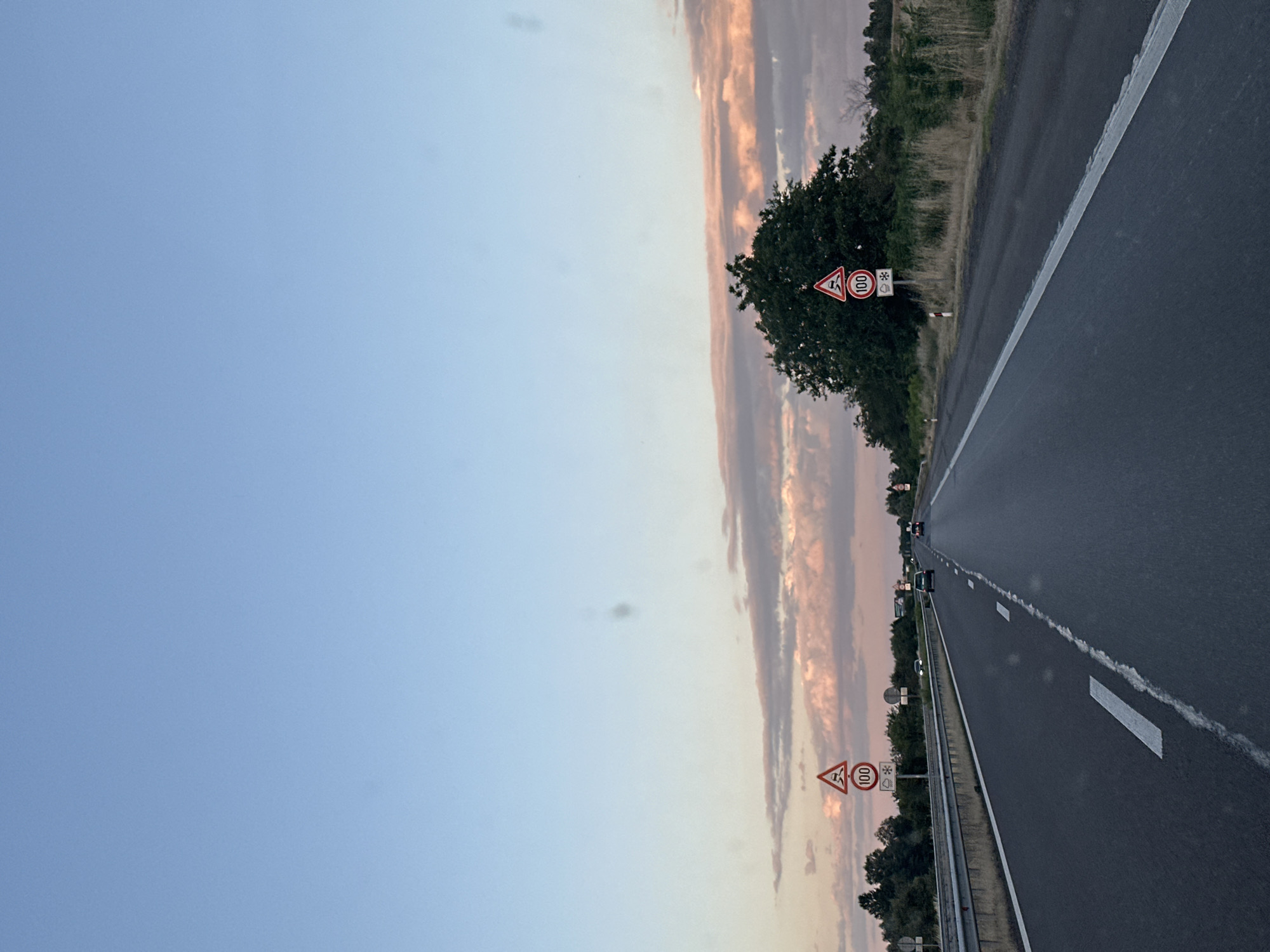 The image shows a view from a vehicle traveling on a highway in Kalugerovo, Bulgaria, likely during sunset. The sky is a clear blue with some wispy pink and orange clouds near the horizon. The road is asphalt with white lane markings. On the right side of the road, there are speed limit signs indicating 100 km/h and a warning for slippery roads. A dense green tree is visible behind the signs. On the left side of the highway, there is a concrete barrier and trees. Several cars are visible in the distance on the highway. The overall scene depicts a typical highway landscape in the late afternoon or early evening.