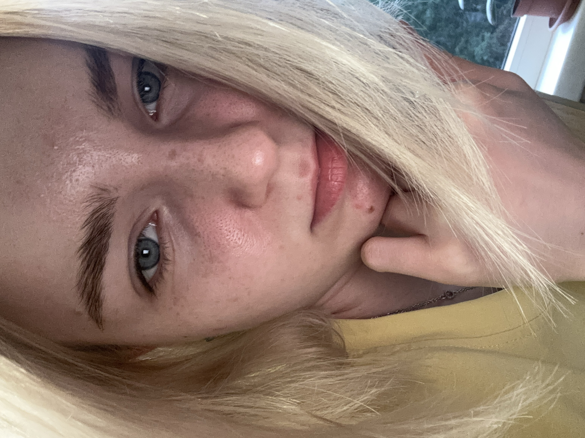 This is a close-up, eye-level selfie of a young Caucasian individual with blonde hair and blue eyes. The individual is wearing a yellow top and a necklace. Their right hand is resting on their chin, and their hair partially covers their face. The background shows a window with a blurry view of trees and a plant. The image appears to have been taken indoors. There is no discernible information about the location in Sinyavino, Russia.