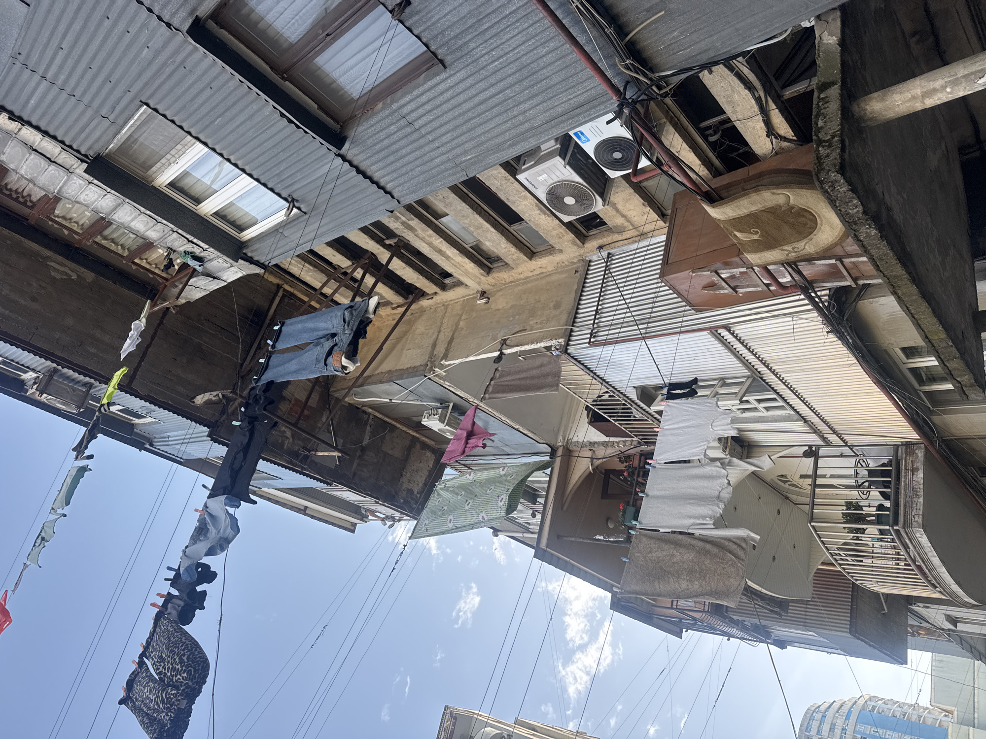 Looking straight up, the image depicts a narrow urban alleyway between buildings, likely a densely populated residential area in the city of Unknown, Unknown. Laundry lines strung between the buildings are laden with clothing and fabric items, including jeans, towels, and sheets. The buildings have corrugated metal roofing and skylights. There is an air conditioning unit mounted on one of the walls. A brown cabinet with a patterned rug is also visible on the side of one of the buildings. A modern building with rounded balconies appears at the edge of the frame. The sky is visible between the buildings, with scattered clouds.