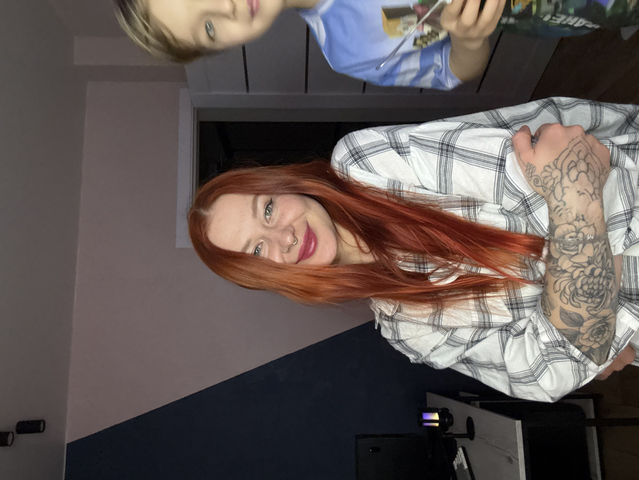 The scene is an interior room, presumed to be in Rostov-on-Don, Russia. In the foreground, an adult female with long, auburn hair, a septum piercing, and visible black-ink floral tattoos on her left forearm, is positioned left-of-center. She is wearing a white and black plaid shirt, smiling directly at the camera. Her left arm is bent across her midsection. To her right, partially visible from the nose downwards, is a juvenile male with light blonde hair, wearing a light blue graphic t-shirt. He holds a thin, reflective object in his left hand. The background wall features a diagonal paint scheme: dark blue on the left, light pink on the right. Overhead, two black cylindrical ceiling lights are visible. A light-colored desk on the left contains a dark monitor, a keyboard, and a small, multi-colored LED lamp. A light-colored doorframe leads to a darker interior on the right, with a segment of a paneled door also visible.