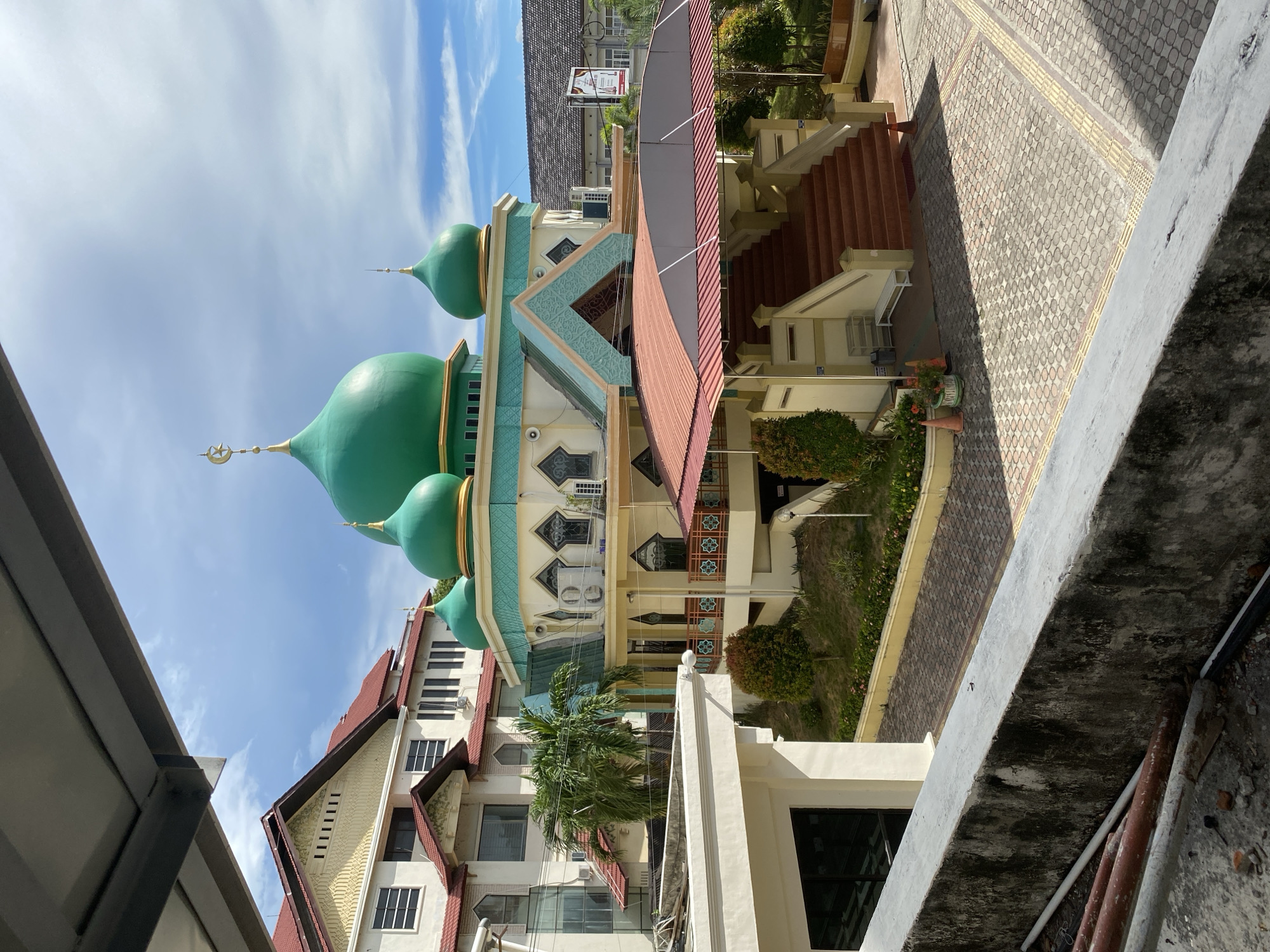 This image shows a mosque with green domes and minarets, located in Banda Aceh, Indonesia. The mosque has a pale yellow facade with teal accents and a red-tiled roof over the entrance staircase. A large building with a red roof and yellow detailing is visible behind the mosque, and a palm tree stands in front of it. The foreground shows a concrete ledge and some construction materials. The sky is partly cloudy.