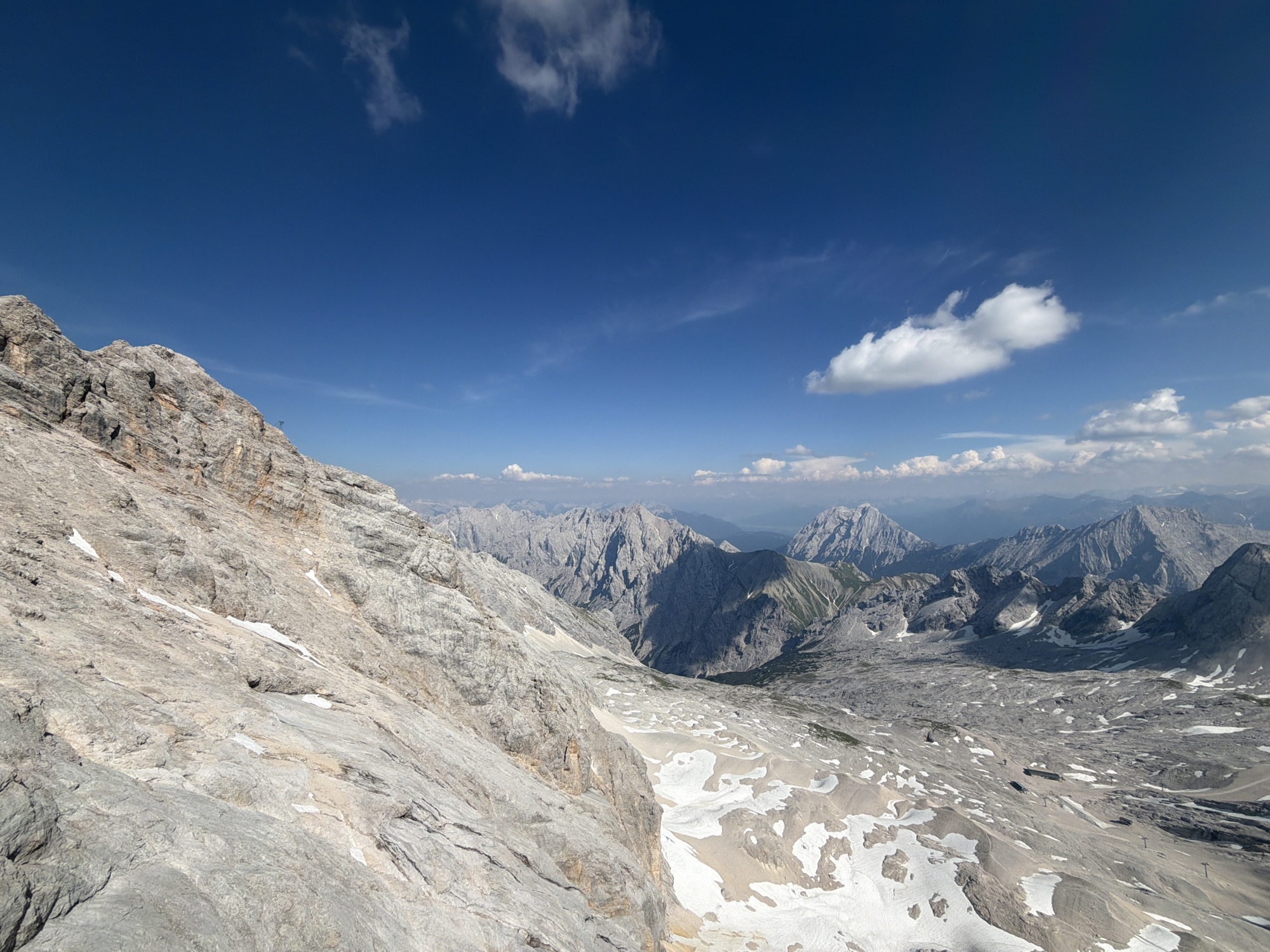 This is a wide-angle, high-angle photograph of a vast mountain landscape under a clear blue sky. The dominant subjects are the rugged, rocky mountains, some capped with snow, stretching as far as the eye can see. The foreground is dominated by a steep, grey, rocky slope, with patches of snow scattered across its surface. Beyond this, the terrain opens up into a valley filled with more mountains of varying sizes and shapes. Some of the distant peaks are shrouded in a light haze.
The sky is a vibrant blue with scattered white clouds, some wispy and high, others more substantial and lower. The lighting suggests it is daytime, likely midday or early afternoon, given the bright sunlight casting shadows on the mountains. The weather appears to be clear and fair, ideal for mountain viewing.
The scene evokes a sense of grandeur, isolation, and the raw beauty of nature. There are no visible people or specific objects like buildings or vehicles, reinforcing the feeling of a remote and untouched wilderness. There is no visible text in the image.
Based on the provided location context of Garmisch-Partenkirchen, Germany, this scene likely depicts the Bavarian Alps, a region known for its dramatic mountain scenery and hiking opportunities. The presence of snow at this elevation suggests it is either early summer, late spring, or autumn, or a permanent snowfield.